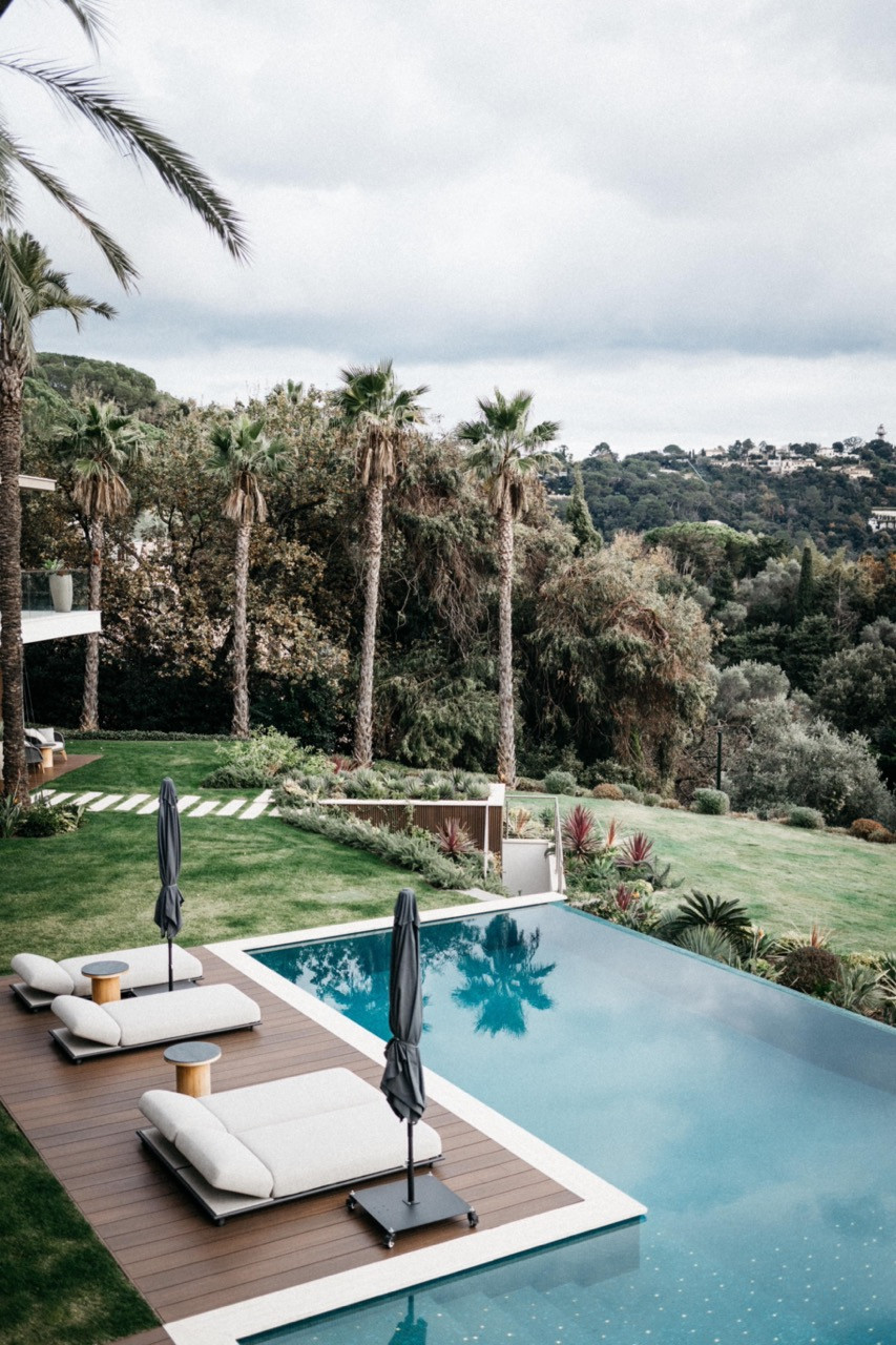 The outdoor swimming pool with upholstered sun loungers on a teak deck, the villa’s two-storey glazed facade and palm trees behind