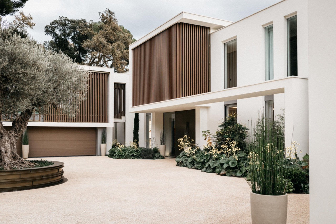 The villa’s entrance forecourt with timber louvred screens, white rendered volumes, a bronze circular planter and established garden planting
