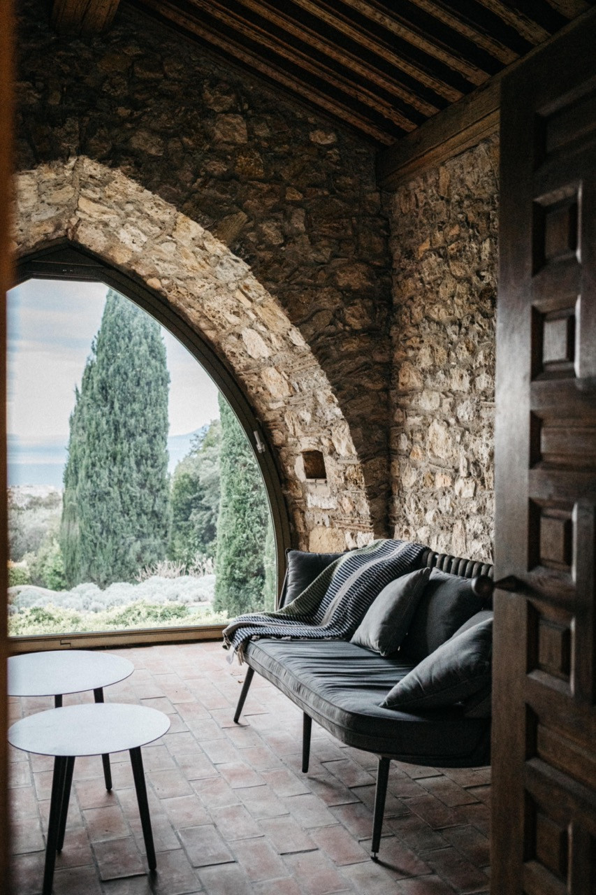 The interior of the restored stone chapel with a pointed stone arch framing views of cypress trees and the Mediterranean, a dark upholstered daybed and terracotta tile flooring
