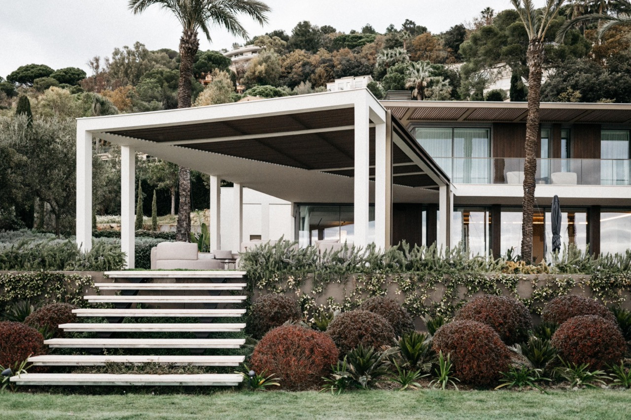 The garden pavilion with a louvred roof, white columns, stone steps leading up from the lawn and layered Mediterranean planting