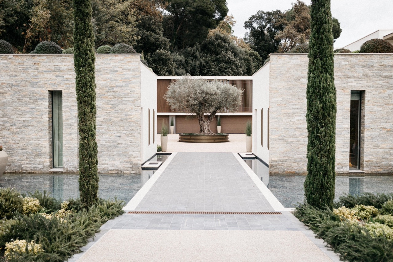 The motor court approach with stone-clad flanking walls, cypress trees, shallow reflecting pools and a mature olive tree in a bronze circular planter