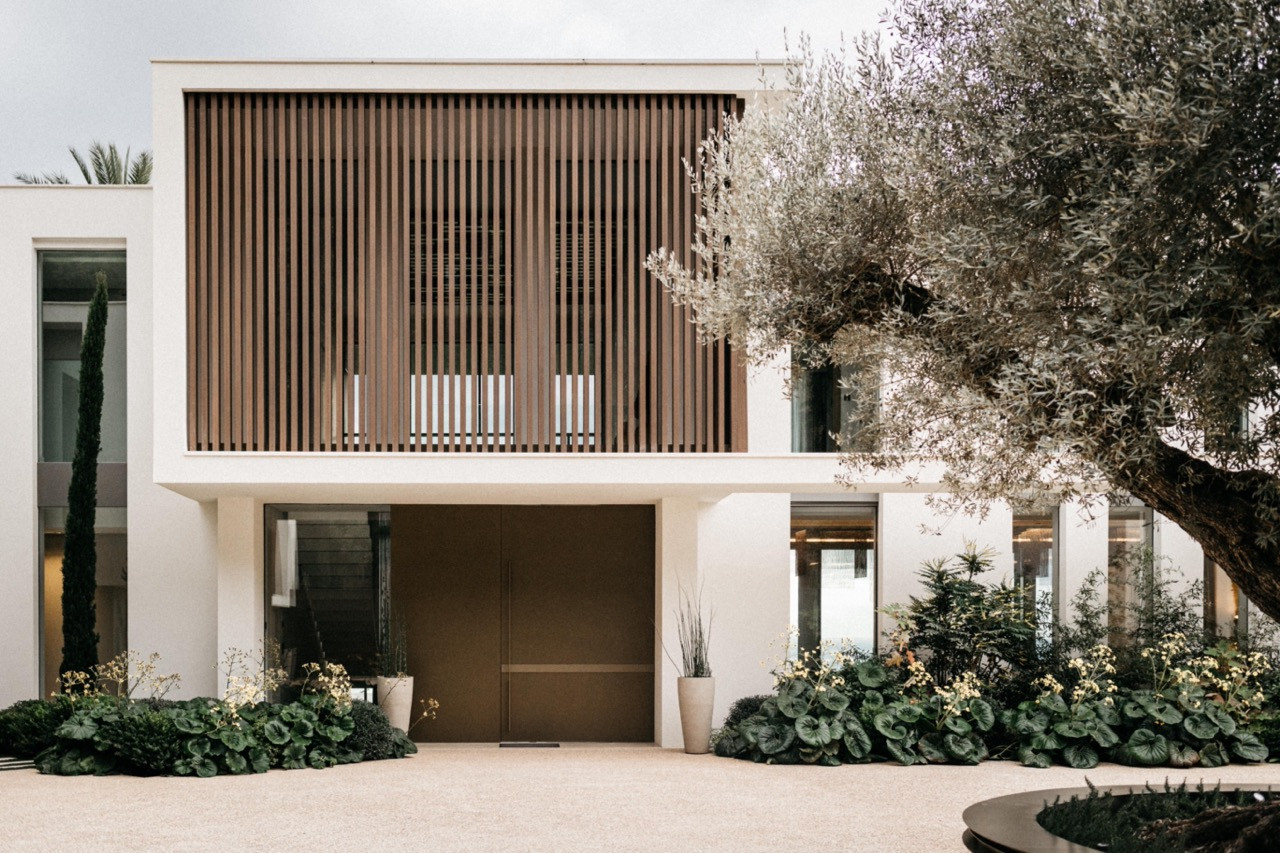 The villa's entrance facade with a large-scale timber louvred screen, white rendered walls, bronze pivot door and established garden planting