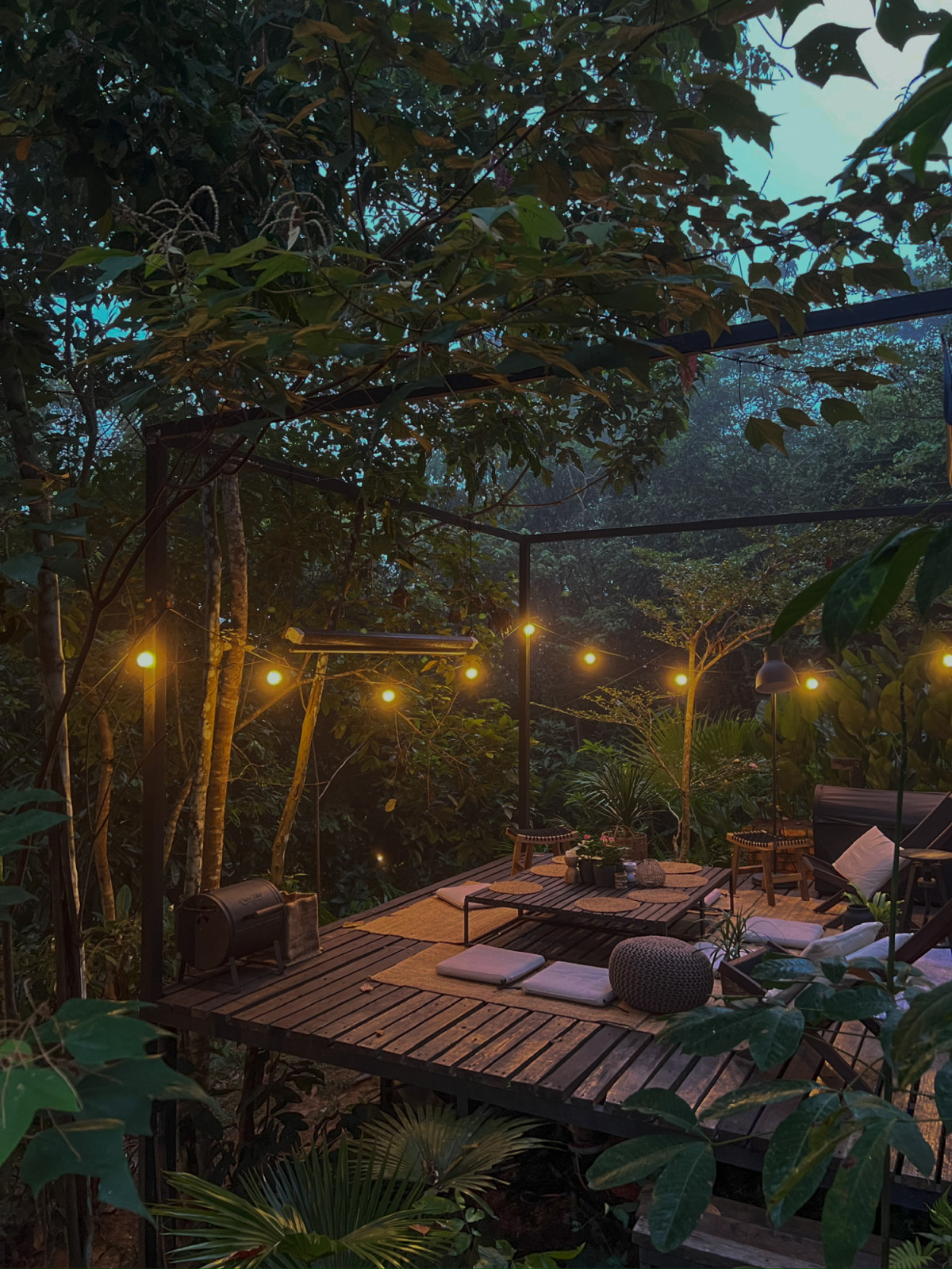 The communal deck at La Hilir lit by string lights at dusk, with low floor seating set against the dense forest understorey