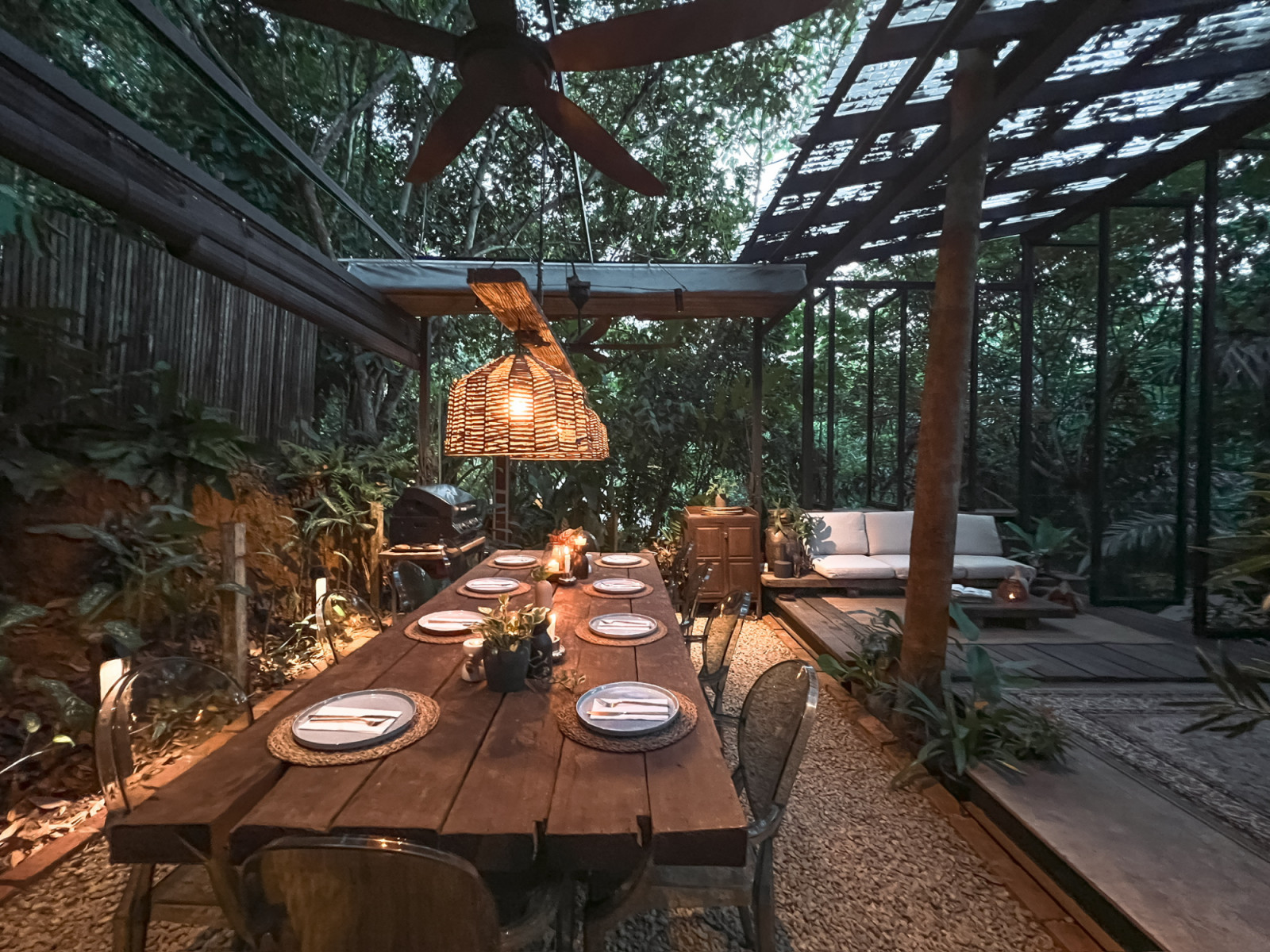 The communal dining and lounge area at La Hilir set for dinner, showing the relationship between the salvaged-timber dining table, rattan pendant light, and the steel-framed pavilion lounge beyond