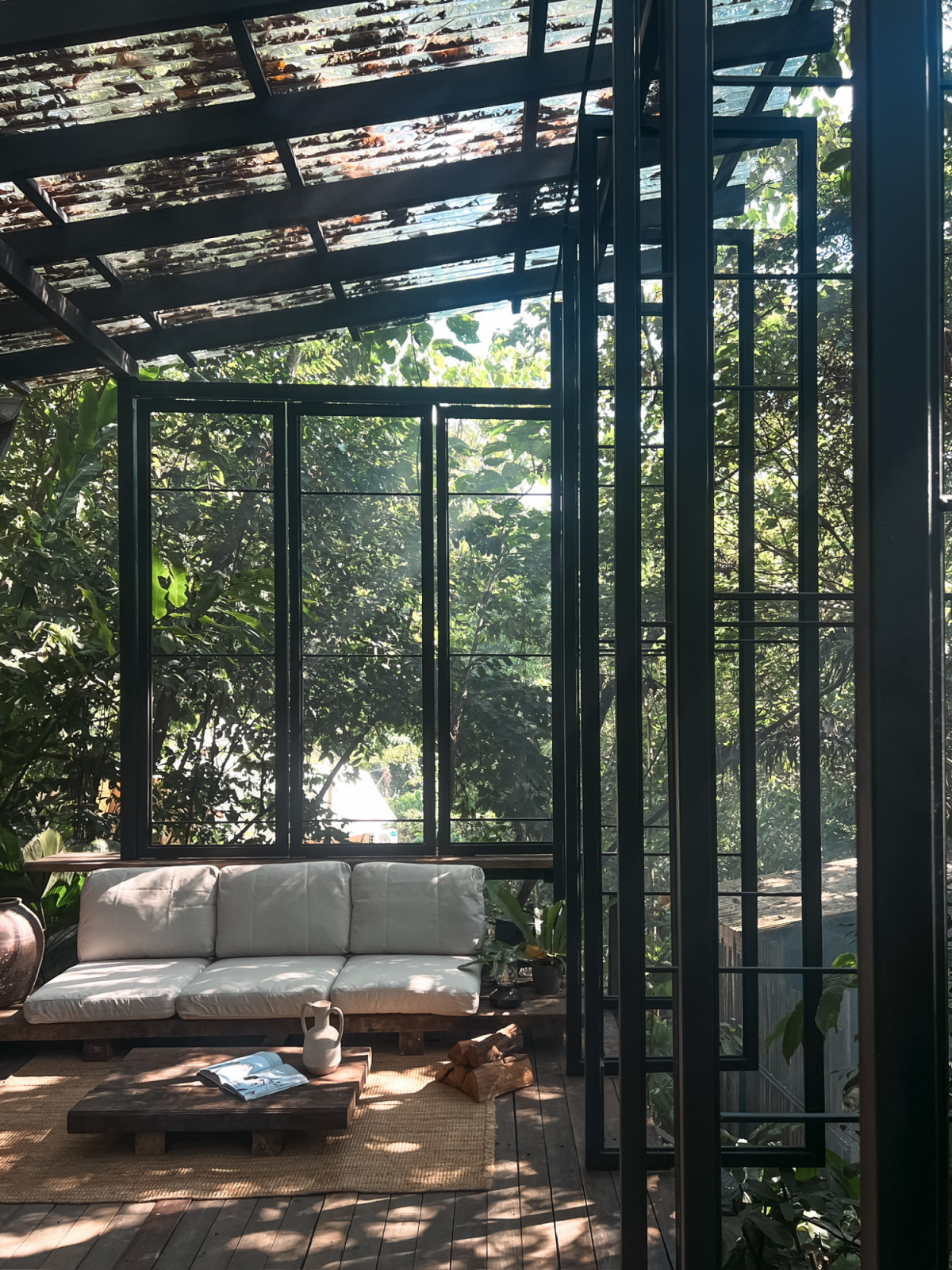The steel-framed pavilion at La Hilir, furnished with a reclaimed-timber sofa base and jute rug, with the forest canopy visible through polycarbonate roof panels