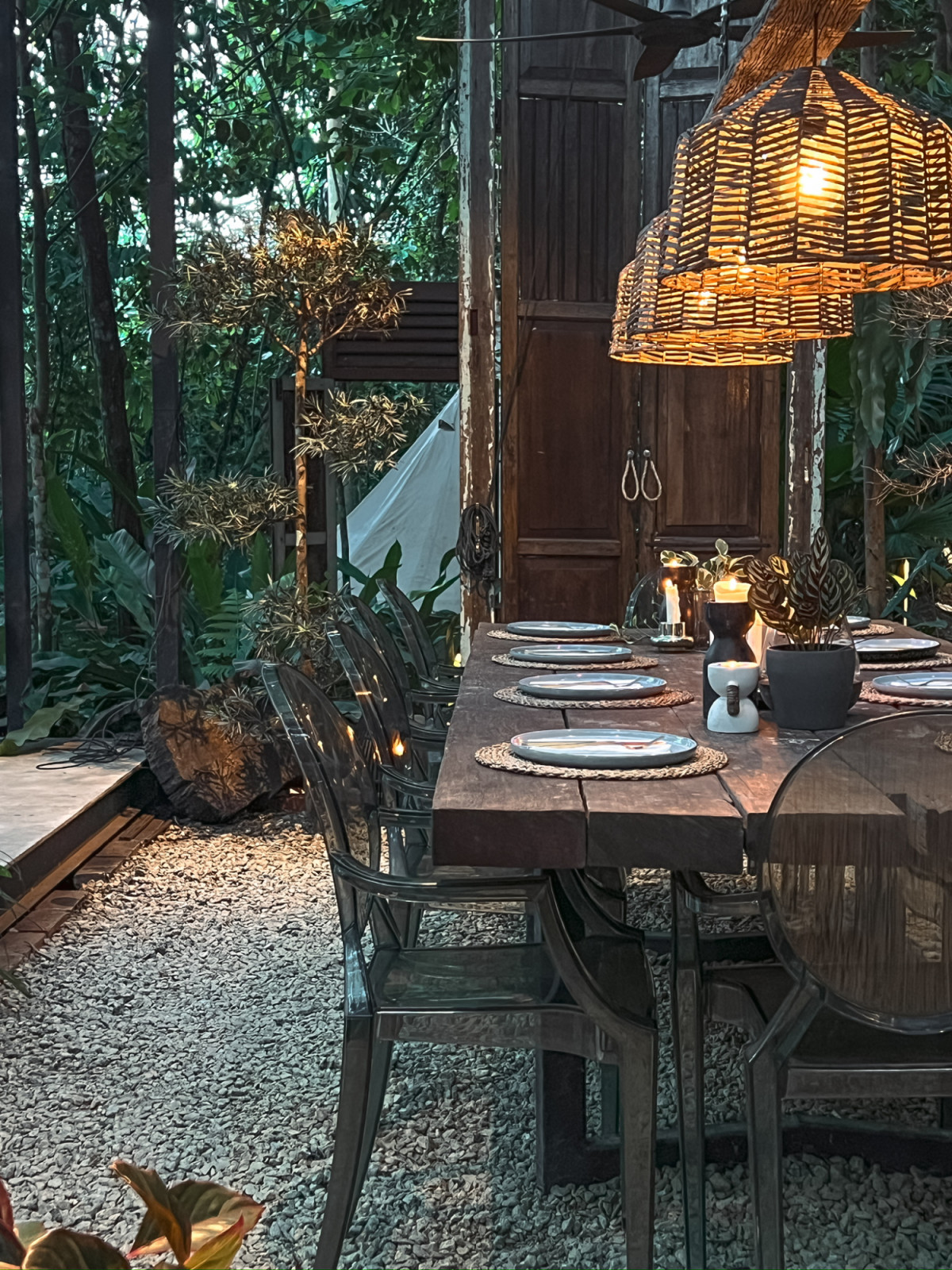The communal dining table at La Hilir set for dinner, built from salvaged kampung house timber and lit by rattan pendant lights, with a canvas tent visible in the background