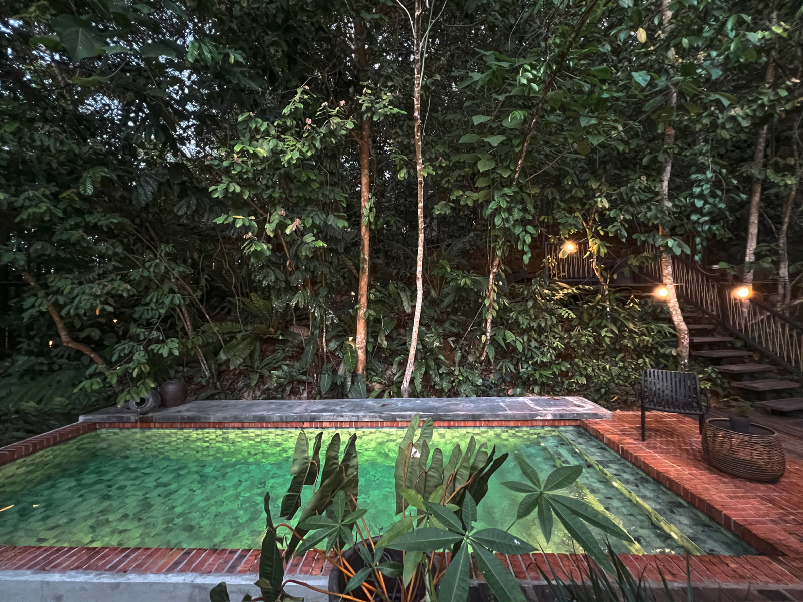 The pool at La Hilir at night, bordered by terracotta brick coping and surrounded by dense forest, with the lit timber staircase to the upper terraces on the right