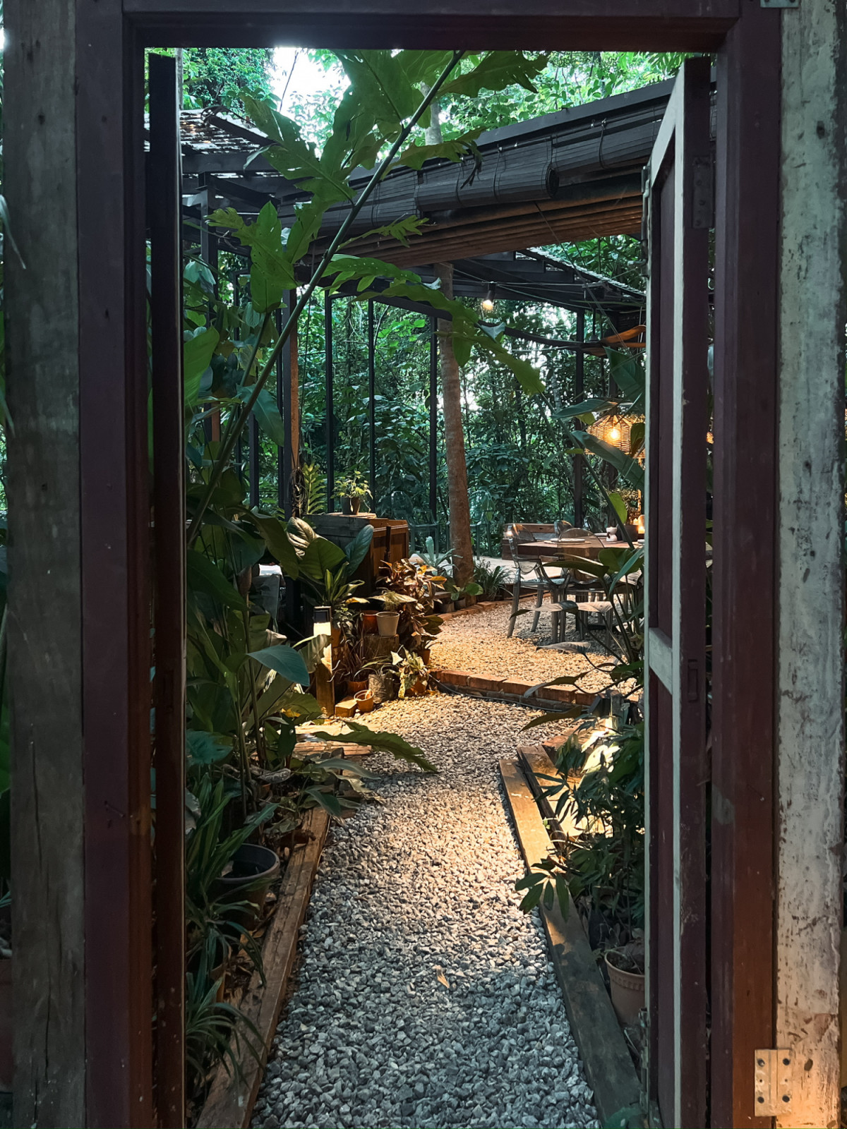 The gravel and brick pathway leading through the site entrance into the communal dining area, framed by a salvaged-timber doorway and dense tropical planting