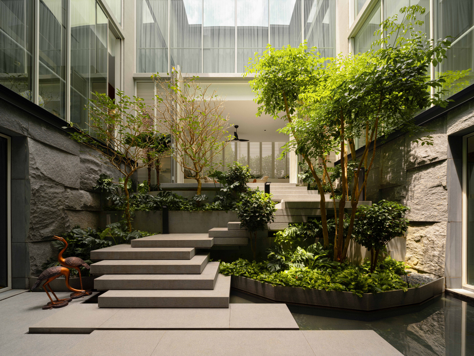 Steps rise through the bungalow’s lush central courtyard, flanked by textured stone walls and dense planting that soften the architecture