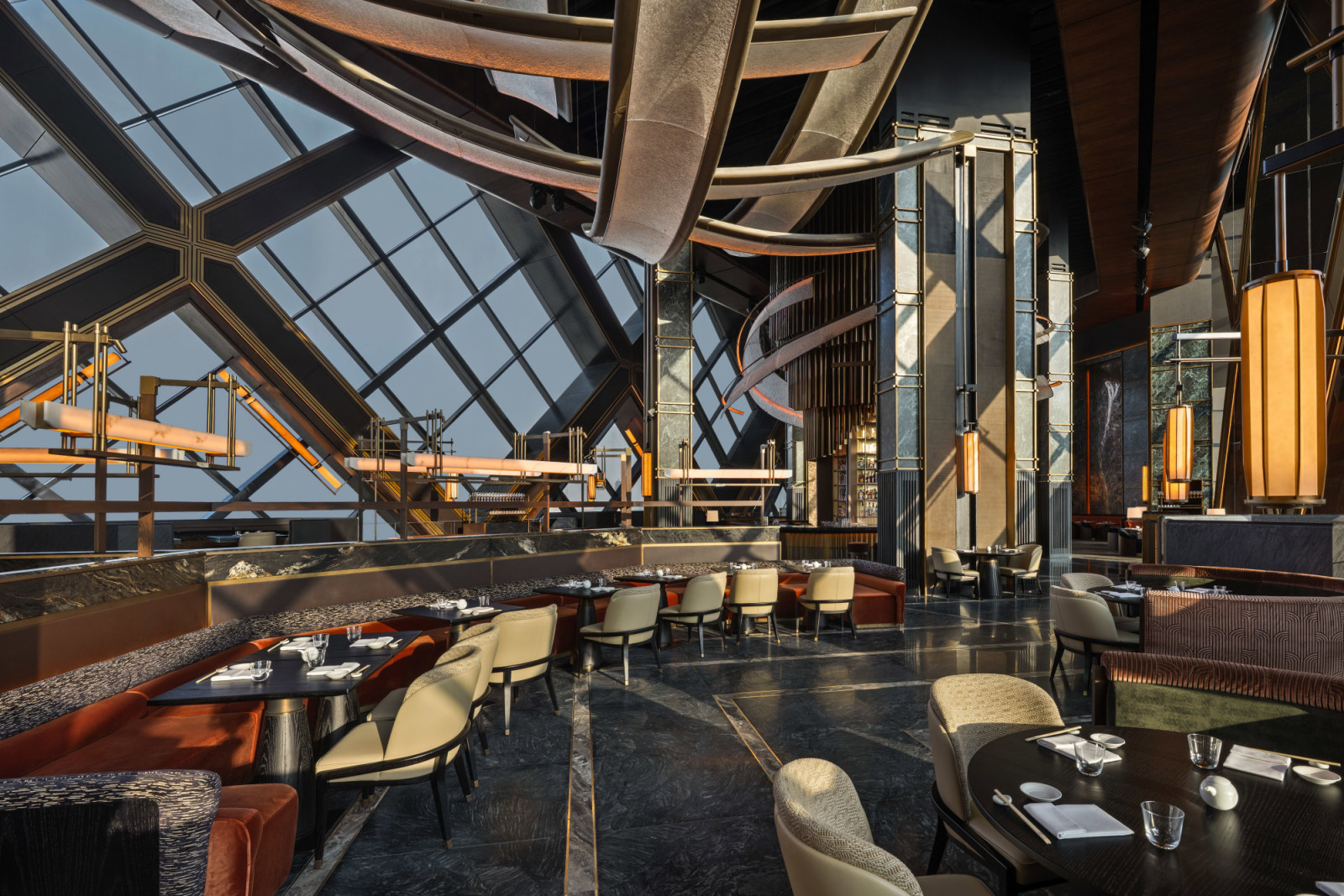 A daytime view of the dining room looking towards the bar, with the One Za’abeel sky concourse structure visible through the angled floor-to-ceiling windows and Rockwell Group’s custom lighting installation overhead (Photo: Alex Jeffries)