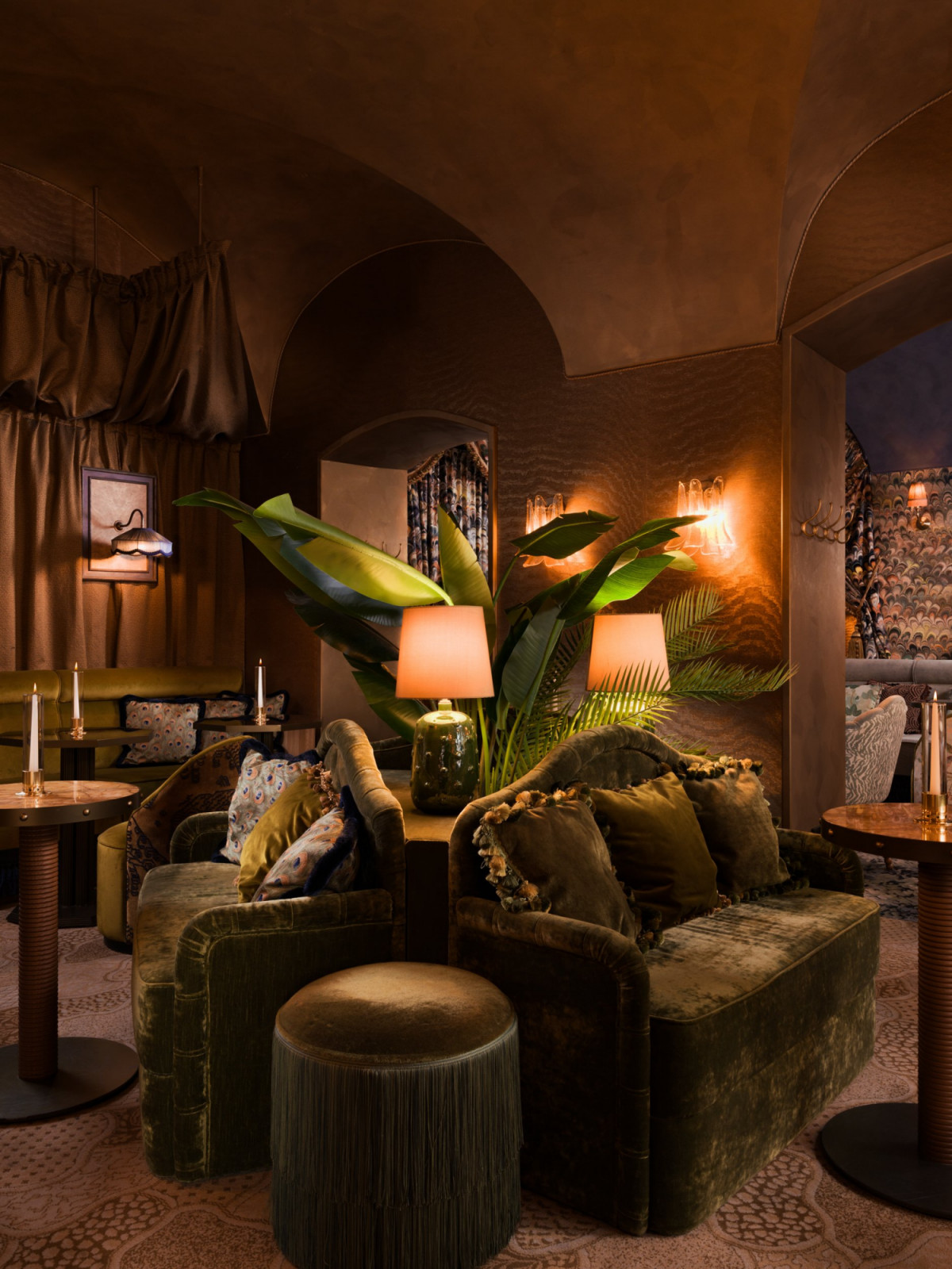 A seating area within Le Fou’s rust and terracotta lounge, furnished with an olive mohair sofa, fringed ottoman and large tropical plants set against a warm plastered arch (Photo: Alex Jeffries)