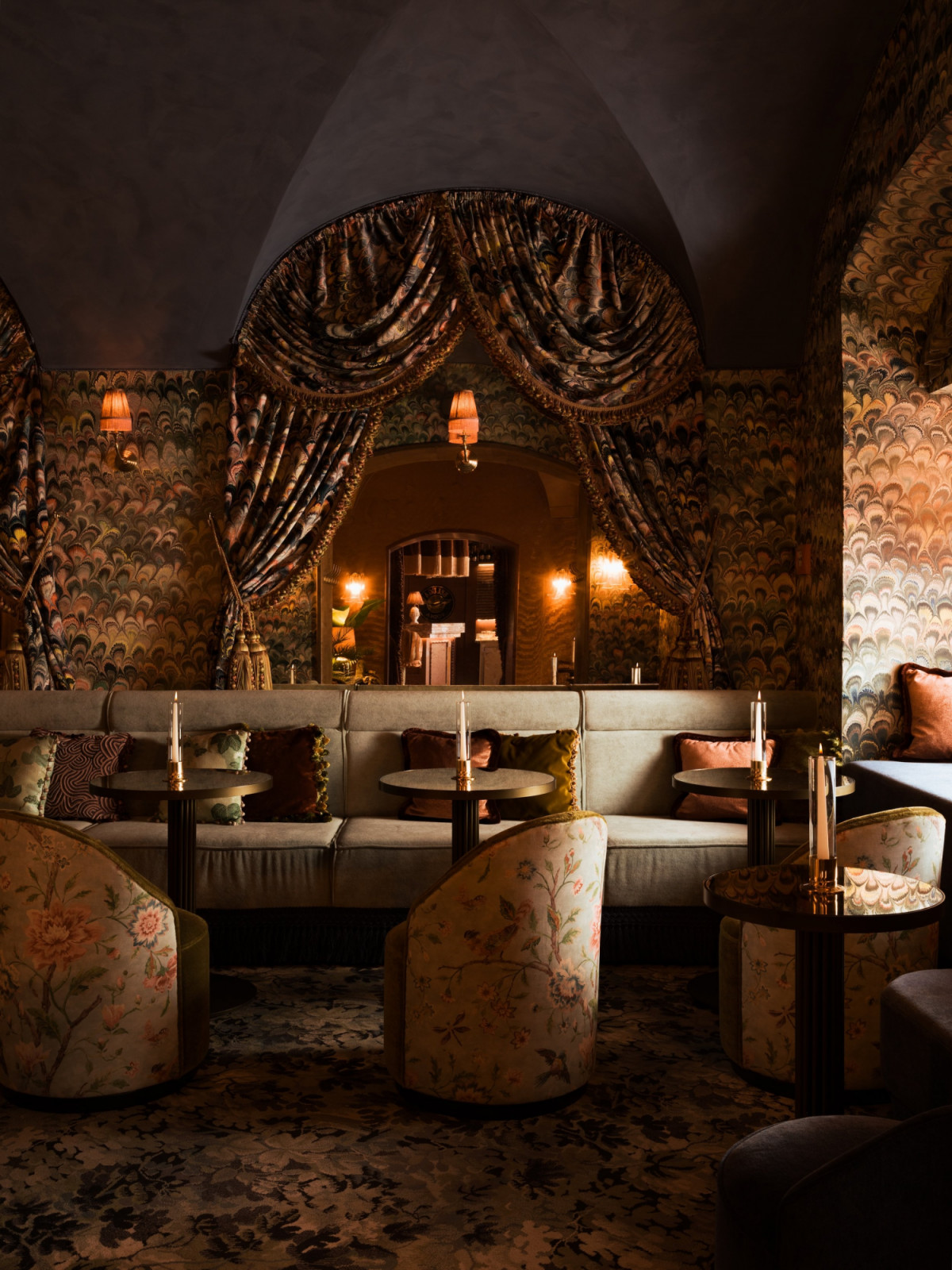 A vaulted lounge room at Le Fou, designed by Theresa Obermoser, with peacock-motif wallpaper, floral barrel chairs and a velvet banquette framed by a draped curtain arch (Photo: Alex Jeffries)