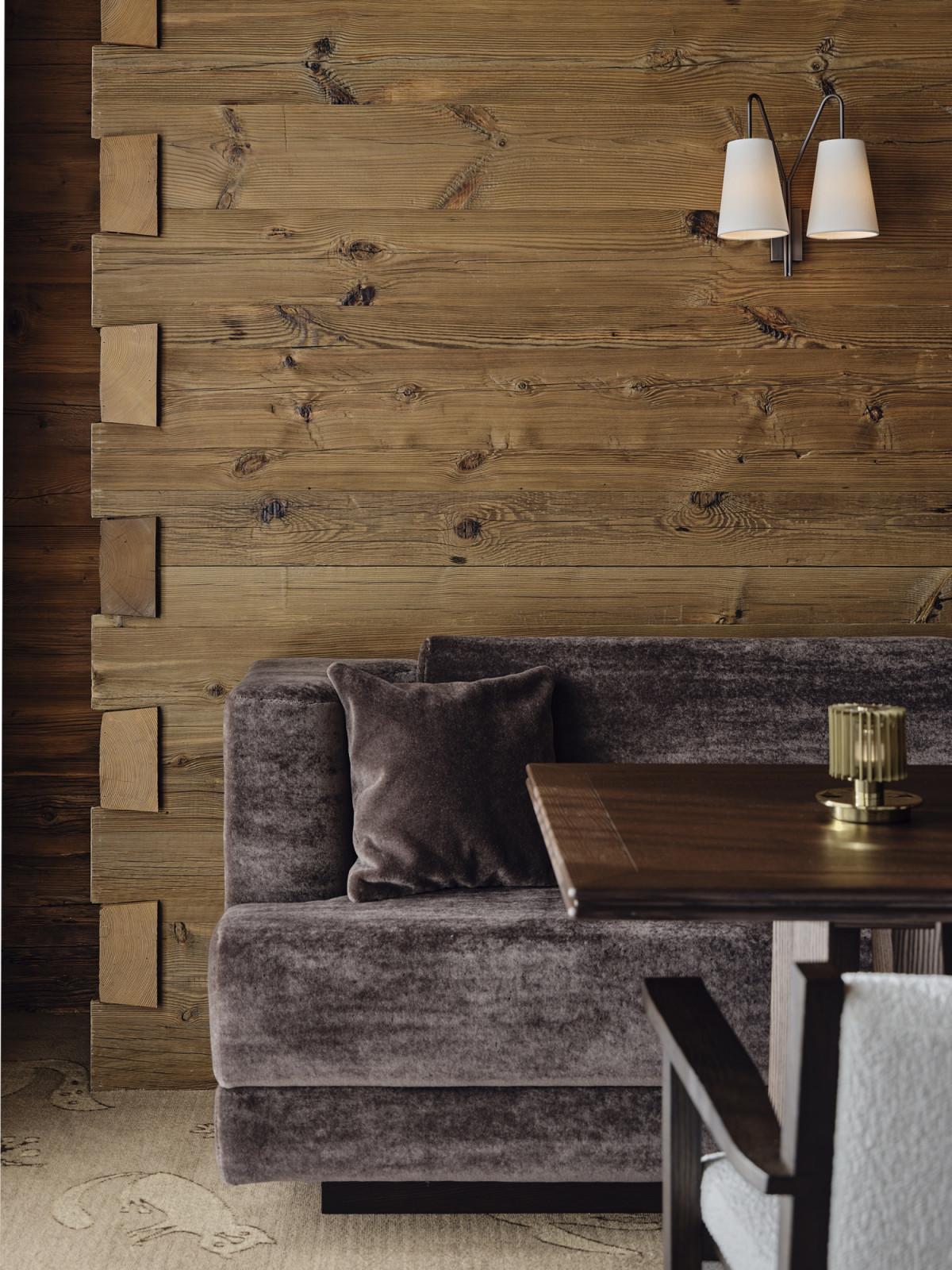 A mohair banquette set against a reclaimed timber wall at Monti Gstaad, with a brass candleholder on the dark wood table and a customised wall sconce above (Photo: Giulio Ghirardi)