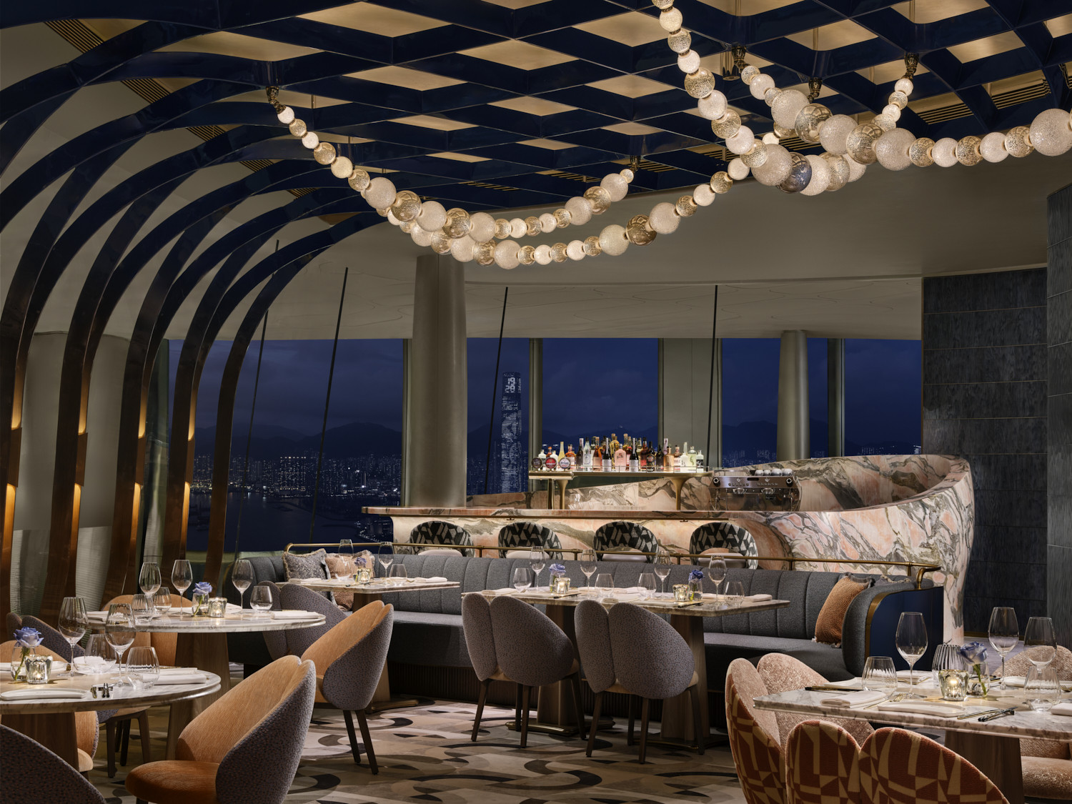 The main dining room at Akira Back Hong Kong, designed by AB Concept, with a curved marble bar, globe lighting installation threading across the diamond-grid ceiling and views of the Victoria Harbour skyline after dark (Photo: Owen Raggett)