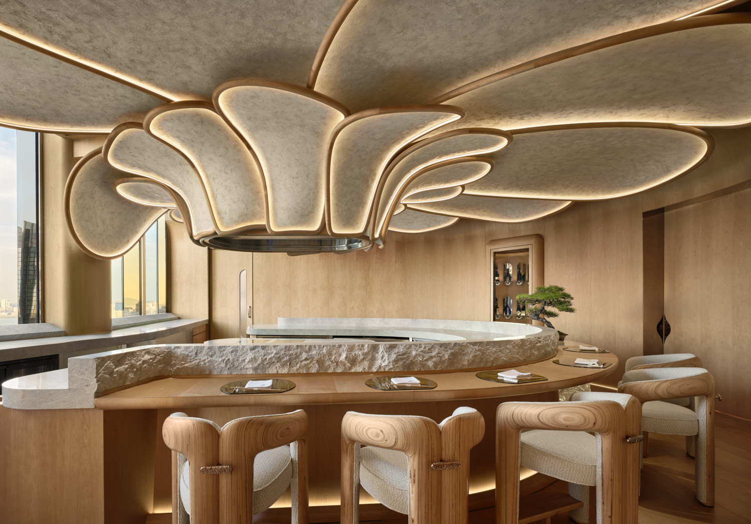 The kappou counter at Hana no Kumo, designed by Hirsch Bedner Associates, with a raw stone surround, cedar bar stools and a petal-form ceiling installation in back-lit plaster above, with a bonsai and views over Hong Kong’s Central district (Photo: Owen Raggett)