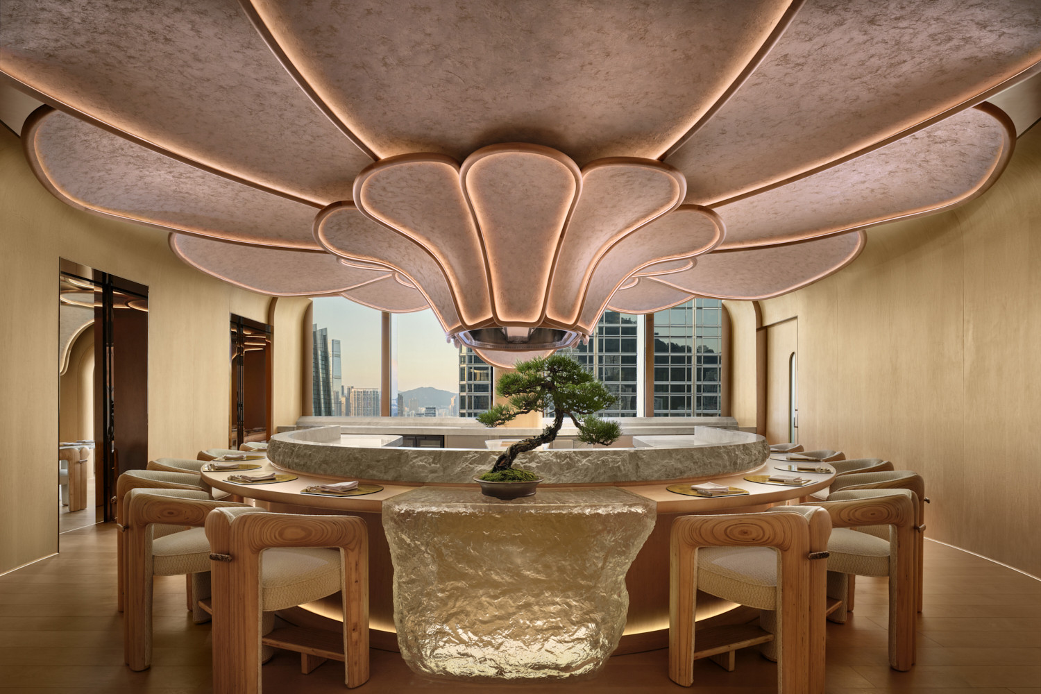 The kappou counter at Hana no Kumo seen from the dining side, with a bonsai at the centre of the raw stone surround, the full petal ceiling illuminated in warm pink tones and views towards Kowloon through the windows (Photo: Owen Raggett)