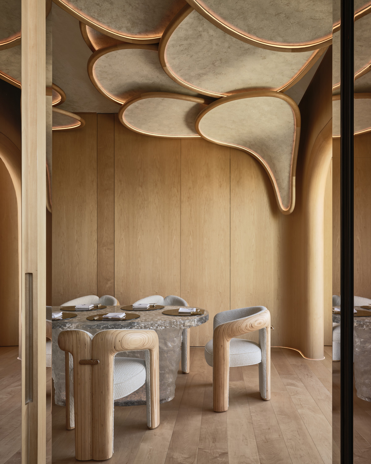 The private dining room at Hana no Kumo, with its biomorphic ceiling panels in pale plaster with brass trim extending over a stone dining table and cedar armchairs, the room reflected in a mirror panel to the right (Photo: Owen Raggett)