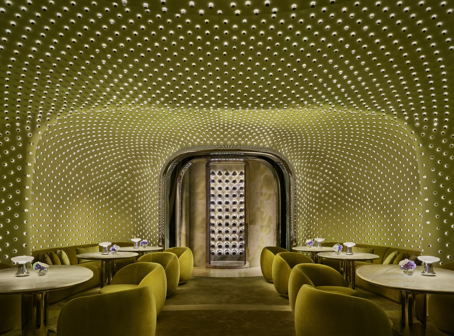A lounge room at Peridot, designed by Studio Paolo Ferrari, with curved walls and ceiling lined with over 20,000 hand-crafted lights in the signature chartreuse palette, with olive velvet armchairs and a mirrored doorway at the far end
