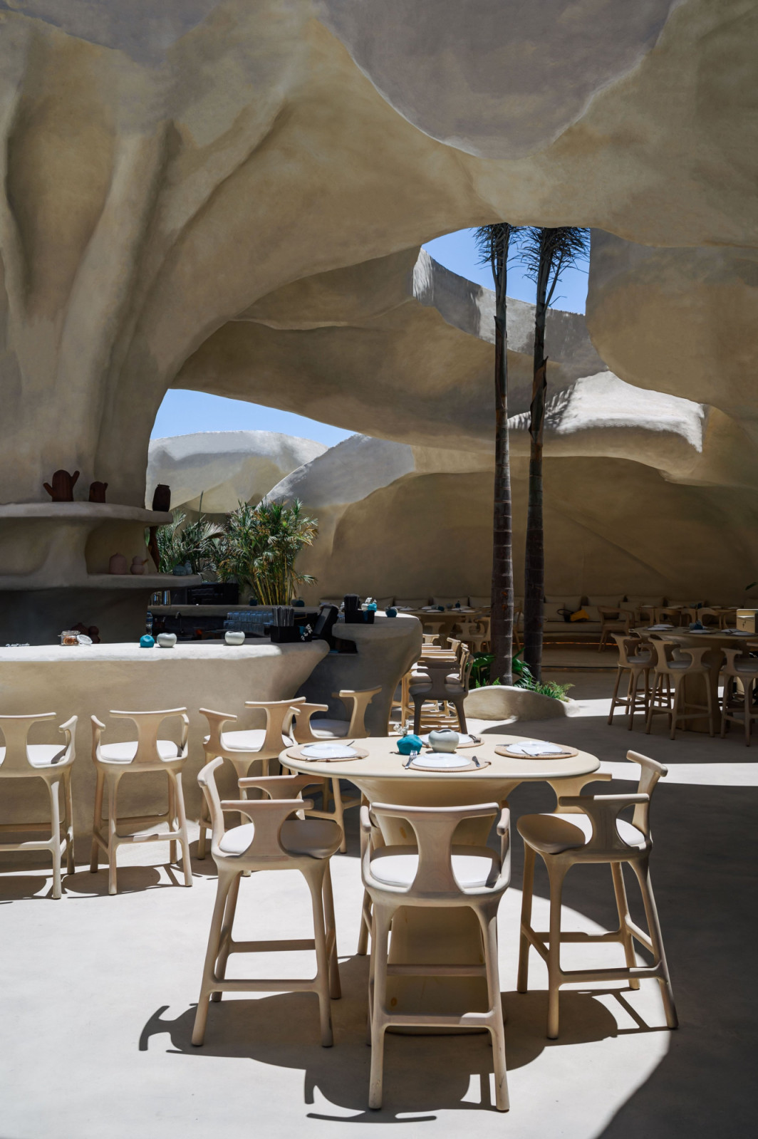 The semi-open dining interior at Escā Playa, its cave-like mineral ceiling pierced by irregular openings to the sky, with bleached wood chairs and tables set around a curved bar, palm trunks rising through the structure (Photo: Nour El Refai)