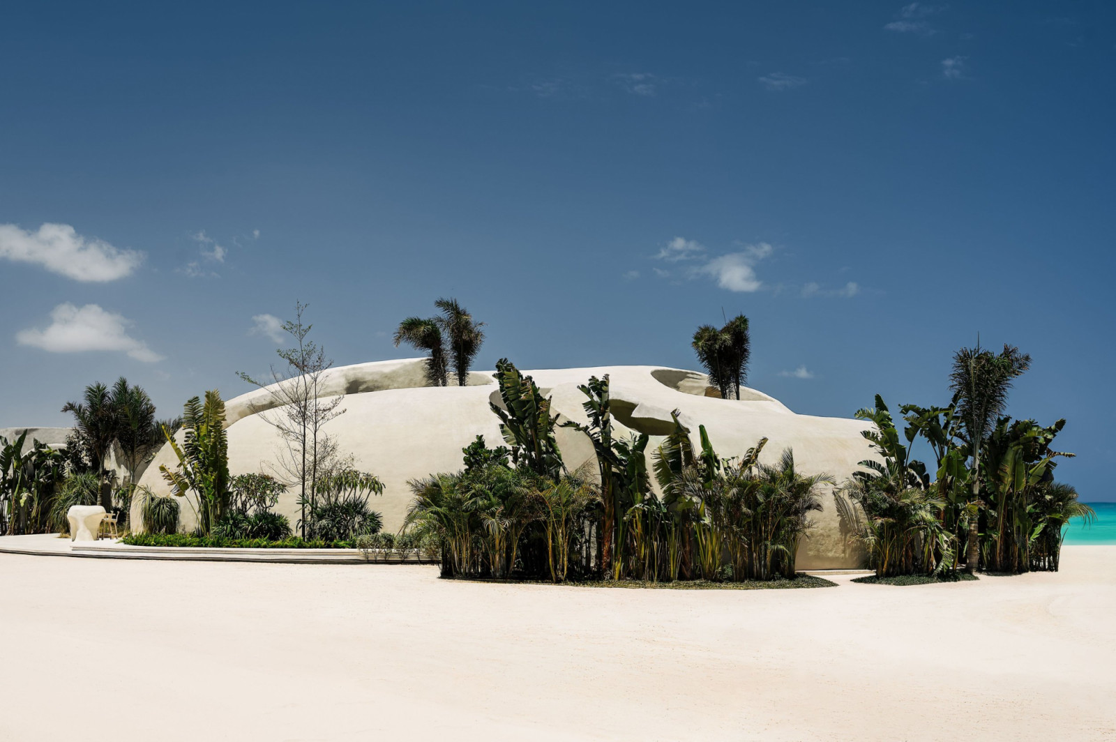 Escā Playa seen from the beach, its low undulating structure embedded in white sand and surrounded by tropical planting, with the turquoise Mediterranean visible beyond (Photo: Nour El Refai)