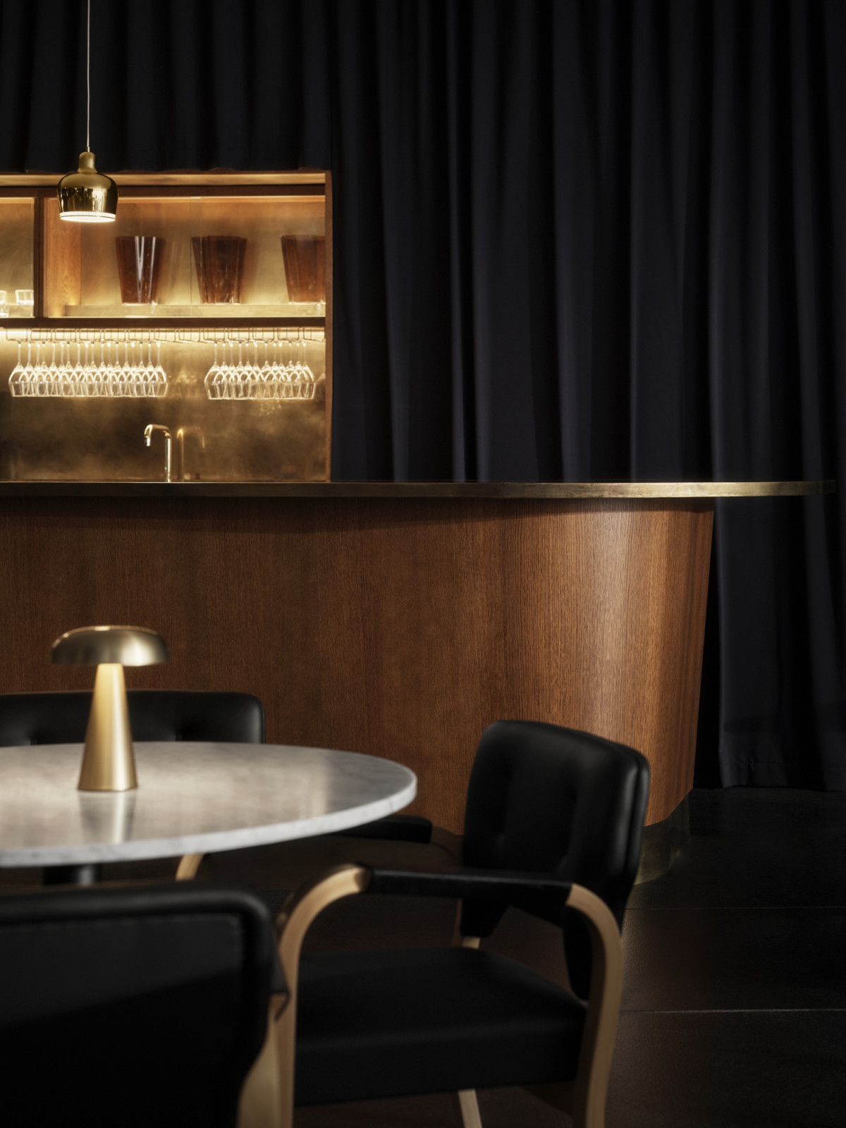 A detail of the bar at Finlandia Bistro showing the brass-lined back shelf, suspended glassware and new pendant lighting above the walnut counter, with Aalto chairs and a marble dining table in the foreground