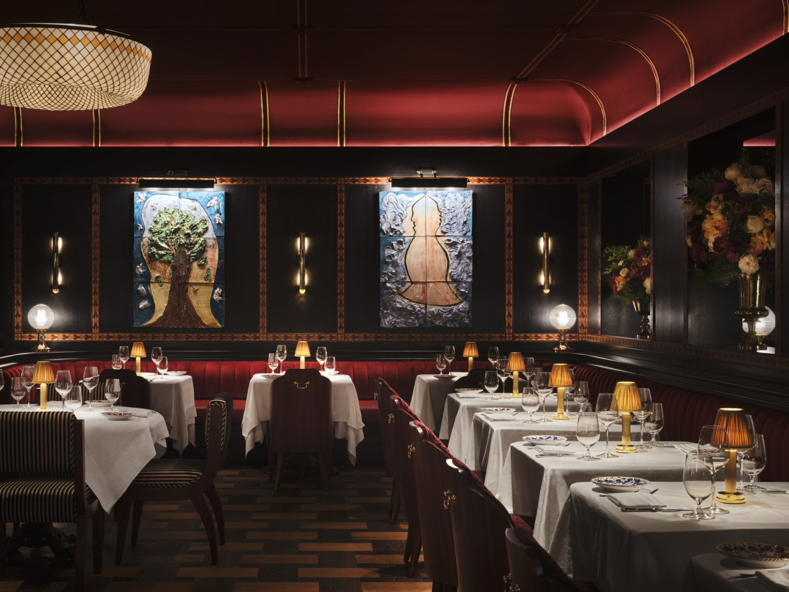 A section of the main dining room at Carbone London showing two ceramic tile relief panels on the dark panelled wall above a crimson velvet banquette, with gold-shaded table lamps and striped dining chairs