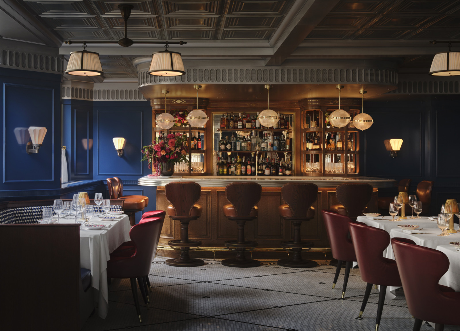 The upper-level bar at Carbone London, designed by Ken Fulk, within the former American Embassy on Grosvenor Square, with a walnut back bar, globe pendants, navy panelled walls and the building’s original coffered ceiling above