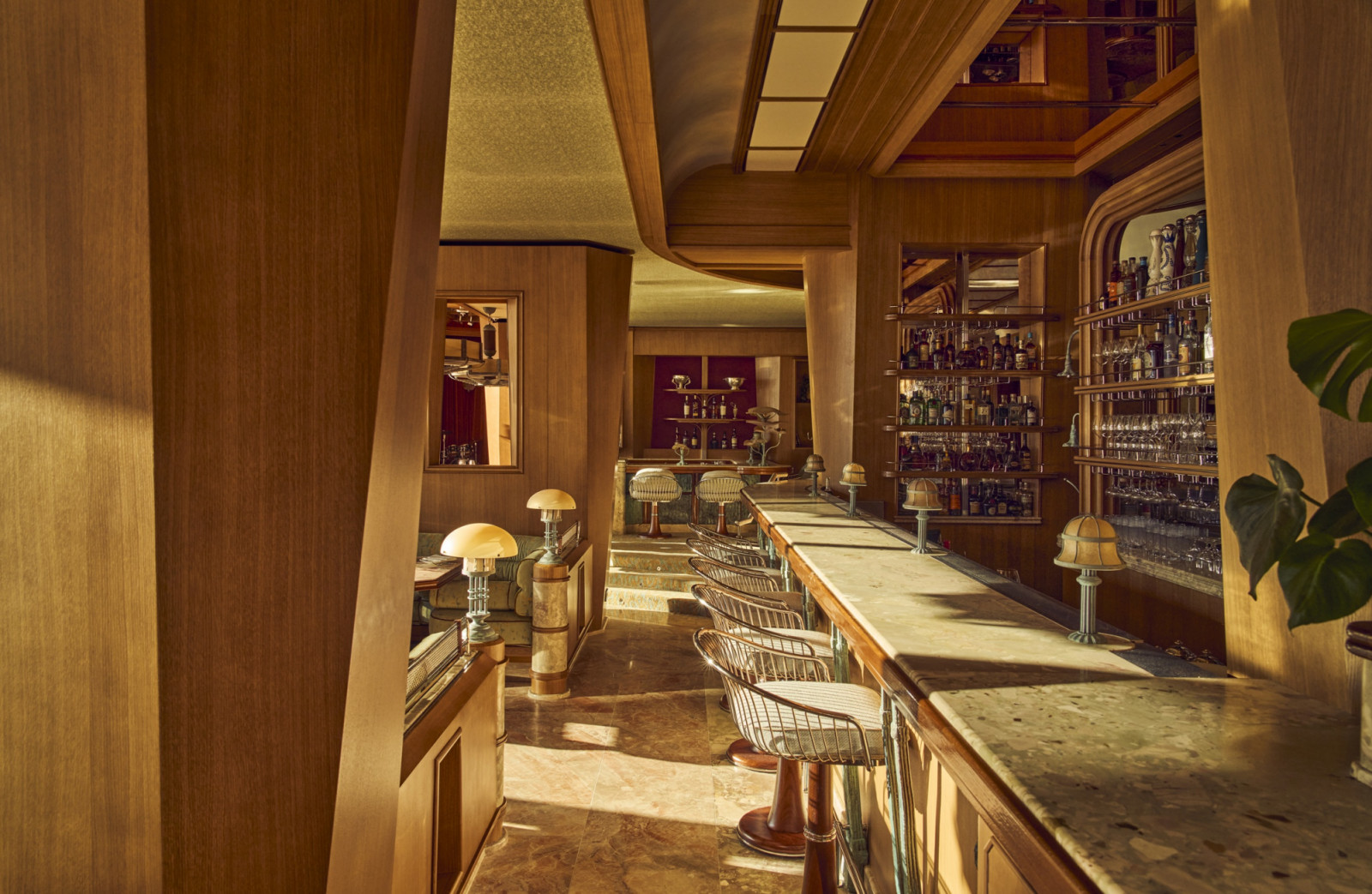 The bar at Marlow, looking towards the dining room beyond, its marble counter lined with wire-frame bar stools and mushroom-cap table lamps, with a full-height bottle and glassware display on the right and warm timber panelling throughout (Photo: Alexandre Tabaste)