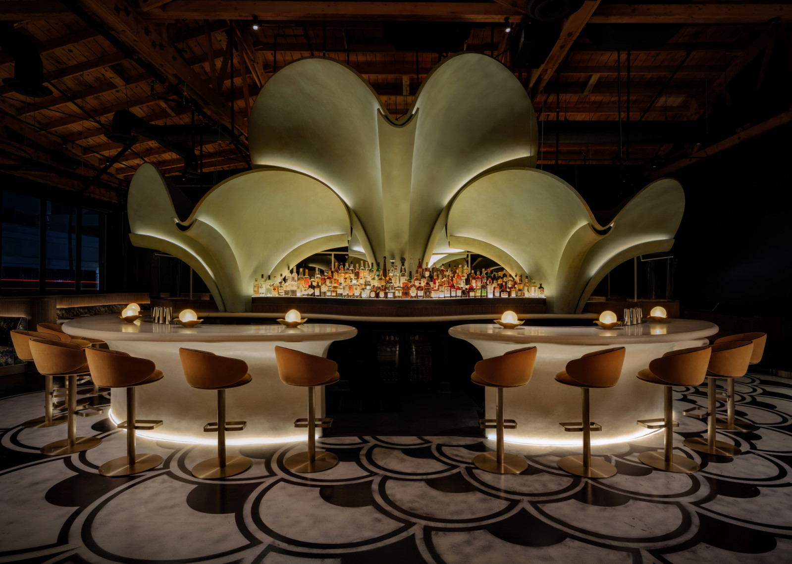 The central bar at Lucia, its oval white counter ringed with brass bar stools and small globe lamps, set against a petal-form sculptural arch backdrop and a black and white scallop-pattern floor