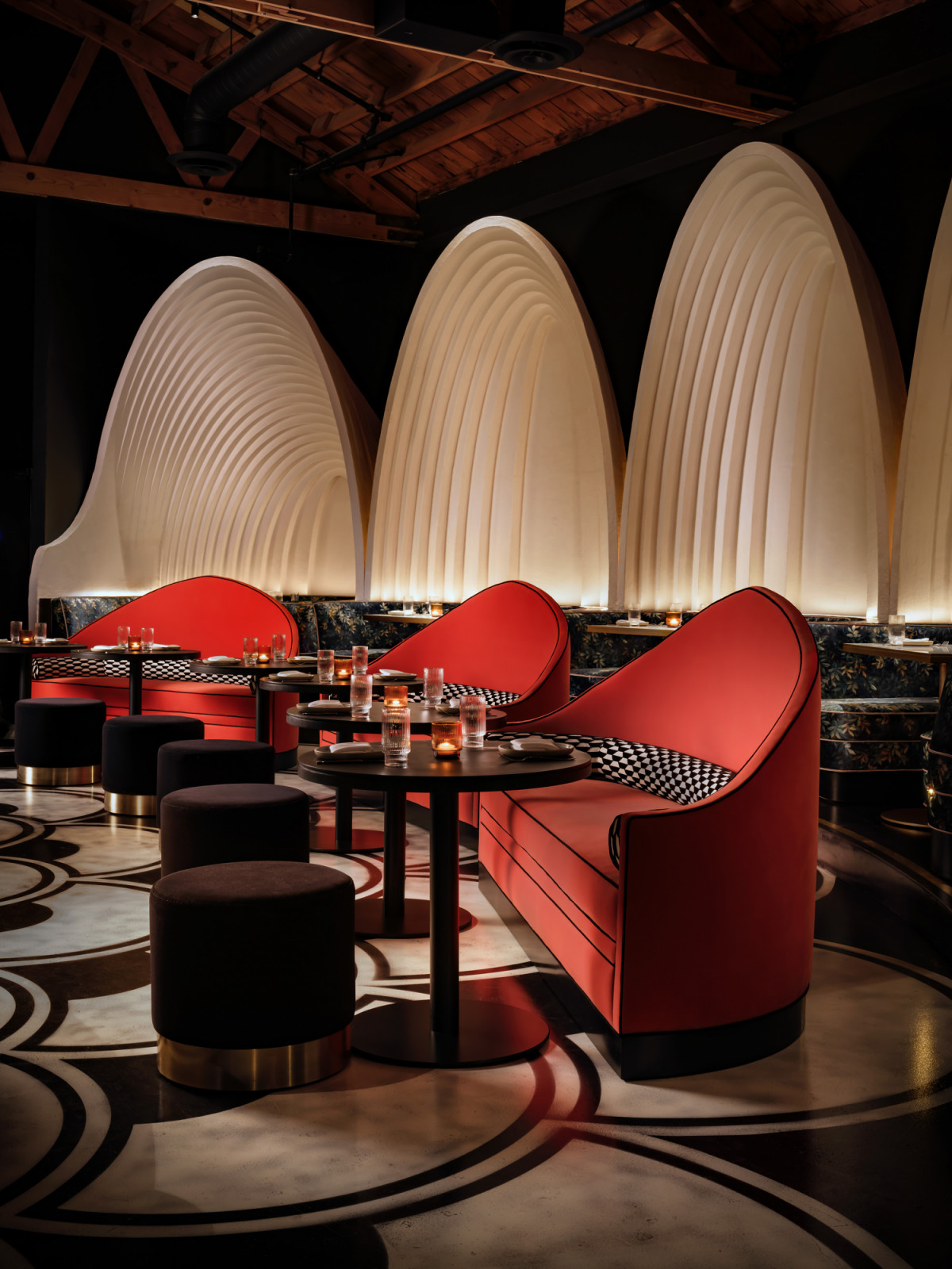 A row of red curved banquettes at Lucia, each set beneath a ribbed plaster arch and upholstered in crimson with a black and white checked trim, with dark ottomans and the graphic floor pattern in the foreground