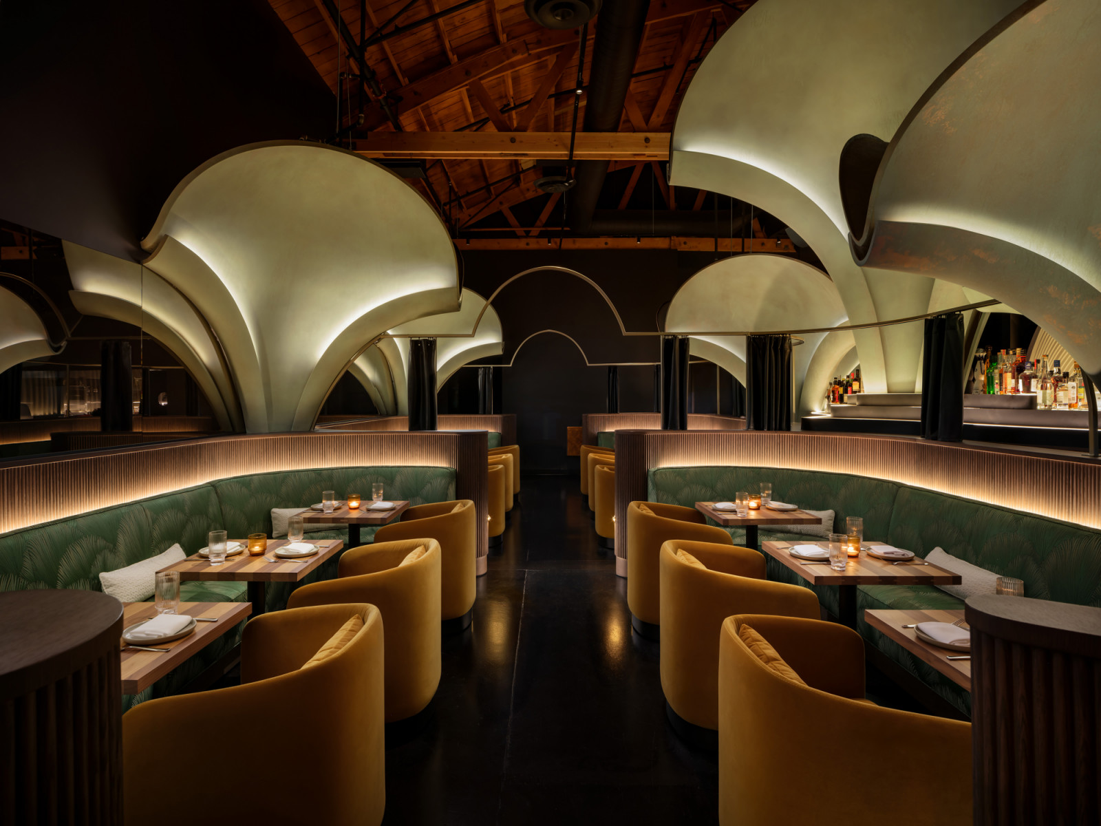 The main dining room at Lucia, Los Angeles, designed by Preen Inc., with back-lit sculptural arch forms overhead, green tropical-print banquettes, gold velvet tub chairs and an exposed timber roof structure above