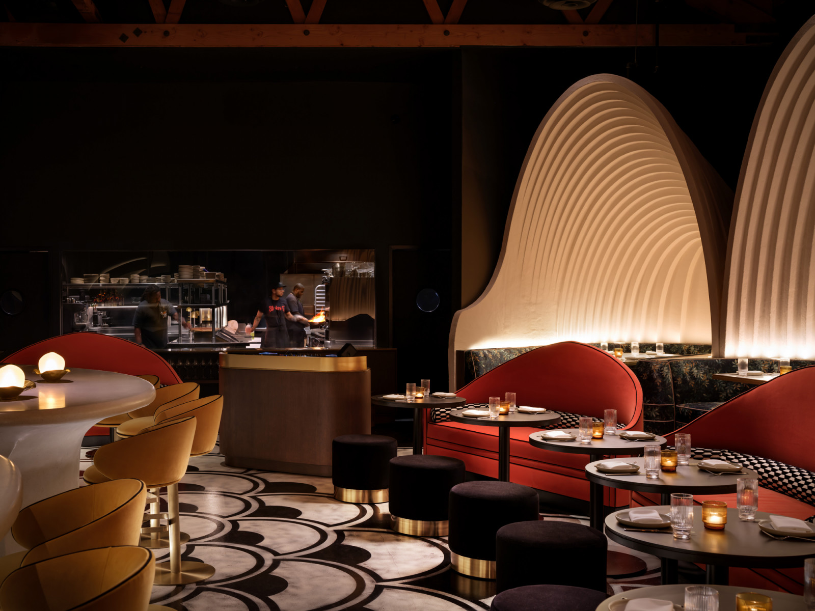 The open kitchen at Lucia is visible through a glass panel, with red curved banquettes set against a ribbed arch surround, black ottomans and the scallop-patterned floor running through to the bar area
