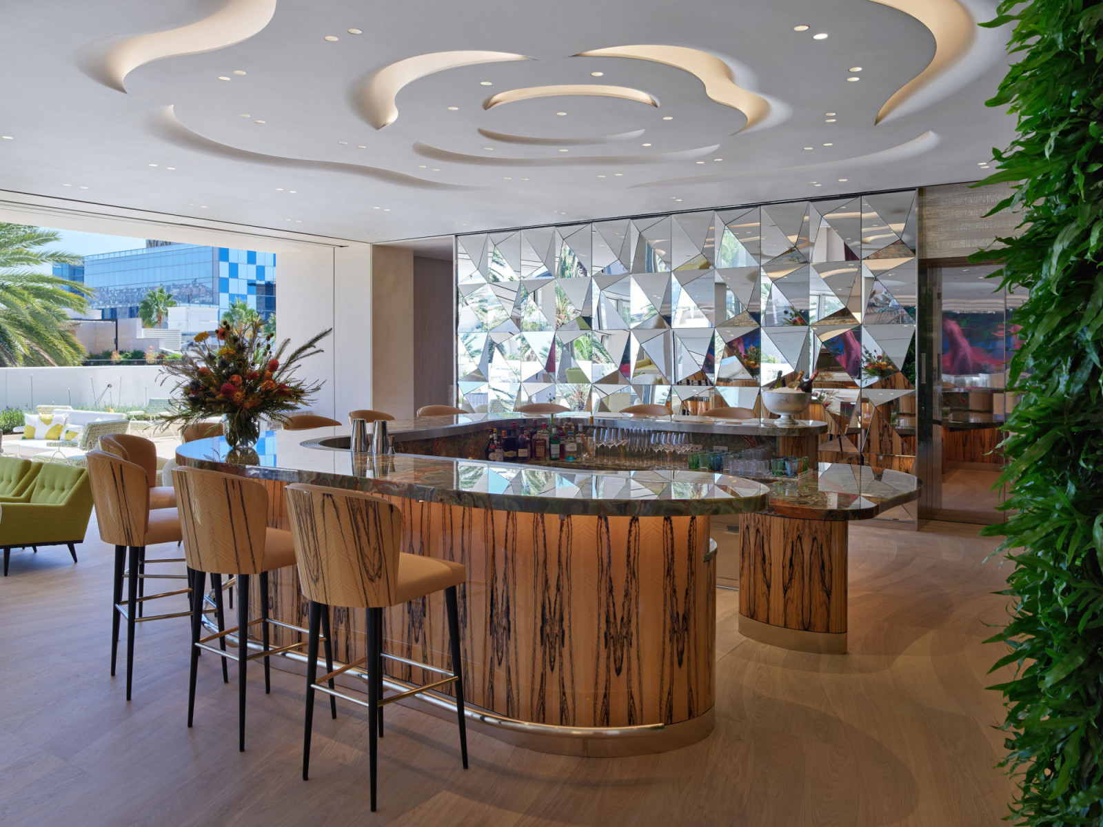 The bar at Monsieur Dior by Dominique Crenn, its ebony and onyx counter set beneath a floral ceiling installation and a faceted mirror wall, with the green living wall and terrace visible beyond