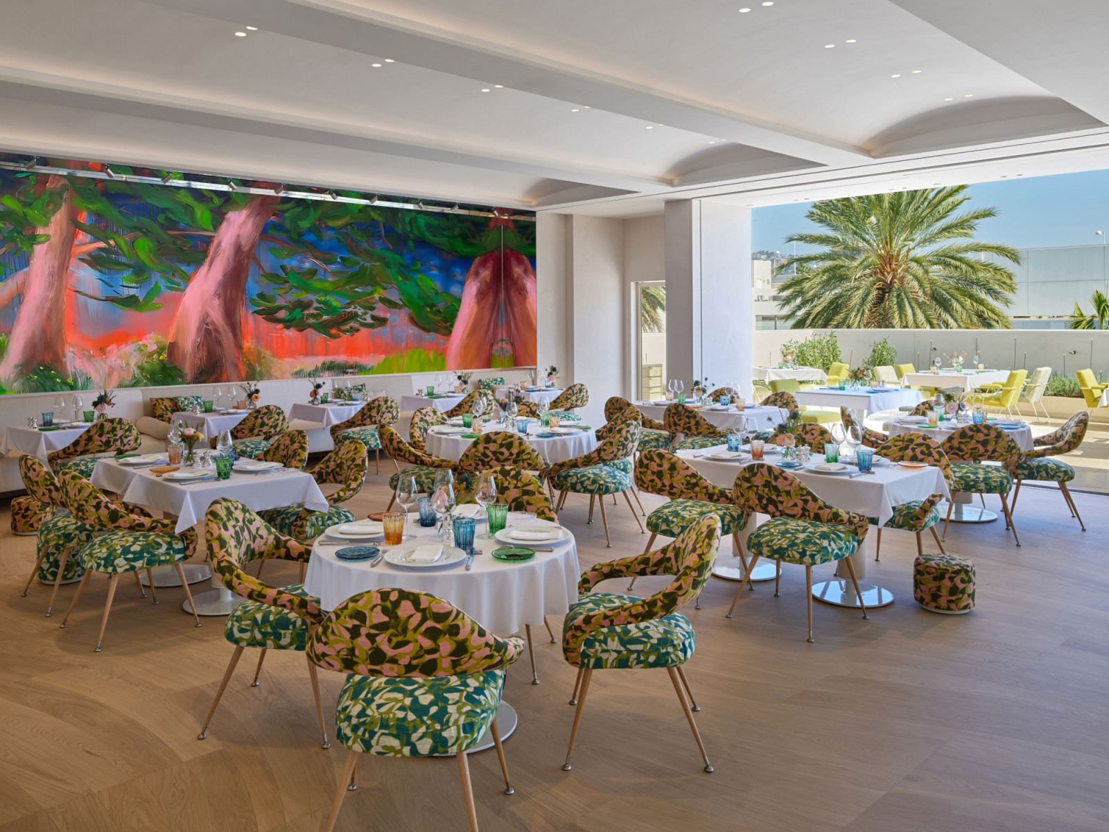 The main dining room showing the monumental Gardens of Courances painting by Nicole Wittenberg on the end wall, with leaf-patterned upholstered chairs in green and pink, white tablecloths and the terrace open to the right