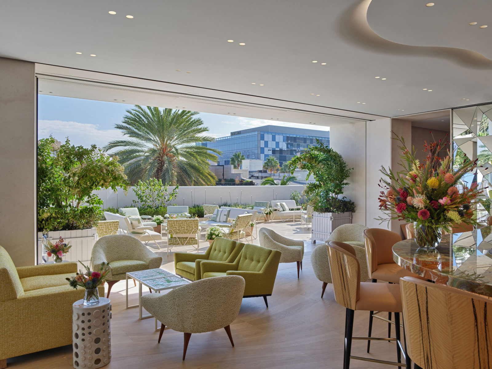 The indoor-outdoor lounge area at Monsieur Dior by Dominique Crenn, with mid-century style green and cream armchairs opening directly onto the terrace garden through a full-height sliding glass wall