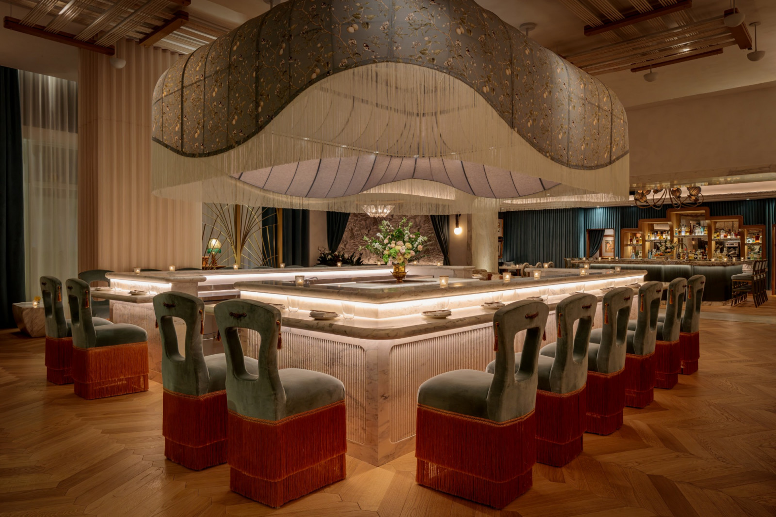 The open sushi bar at Mottai, its rectangular marble counter ringed with green velvet and terracotta-fringed counter chairs beneath a large sculptural canopy of draped fabric and fringing