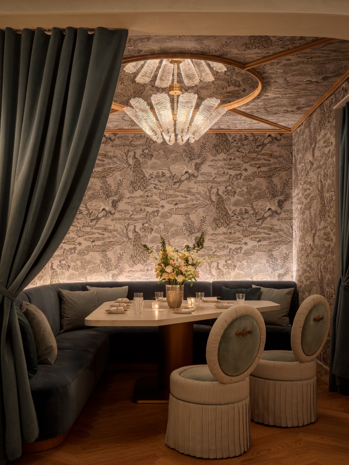 A private dining nook at Mottai, Coral Gables, designed by Saladino Design Studios, with peacock-motif wallpaper running from ceiling to floor, a steel-grey velvet banquette, a fringed chandelier and circular ribbed dining chairs