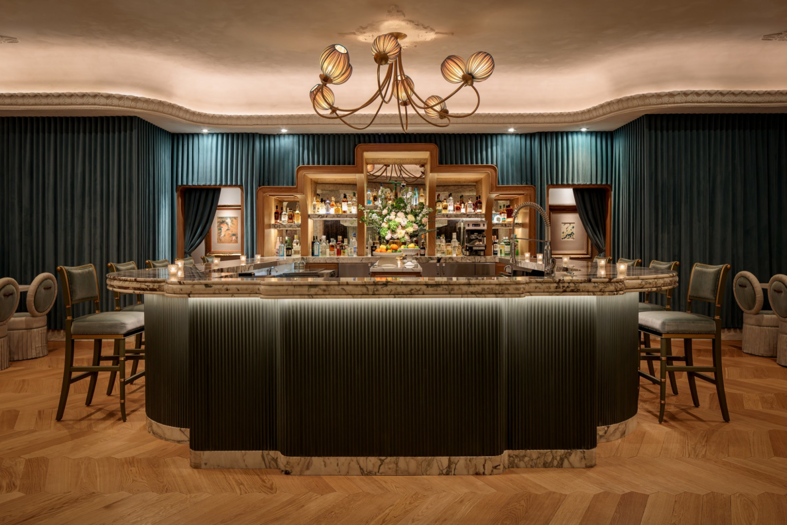 A full view of the cocktail bar at Mottai, its dark fluted counter and marble top set beneath a bronze multi-arm chandelier with pleated shade globes, flanked by teal curtaining and oval-backed bar chairs on either side