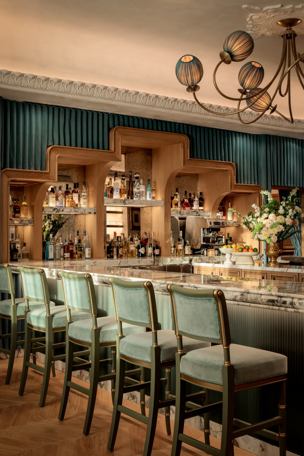 The cocktail bar at Mottai, its marble counter set against a timber arched back bar with teal pleated curtaining, sage green and gold bar stools and an ornate plaster cornice above