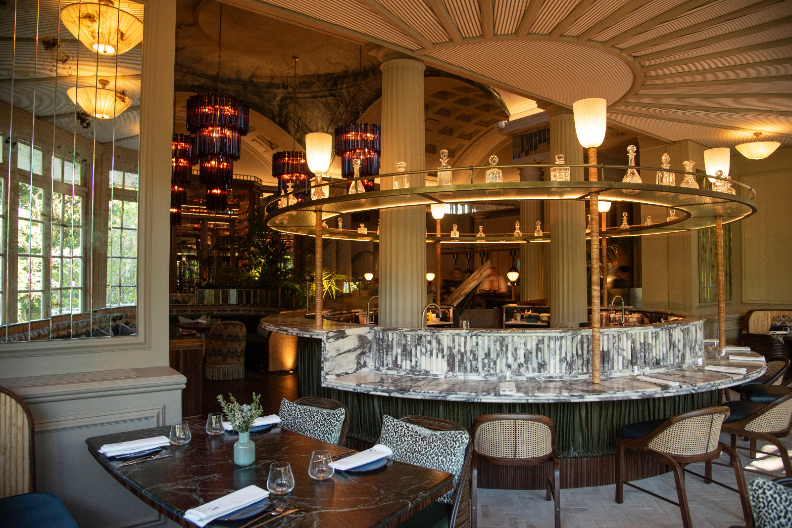 The circular open kitchen at Amura, its marble-clad counter ringed by a brass halo display shelf holding crystal decanters, with tiered amber glass chandeliers beyond and the restaurant’s vaulted classical architecture visible throughout (Photo: Claire Gunn)