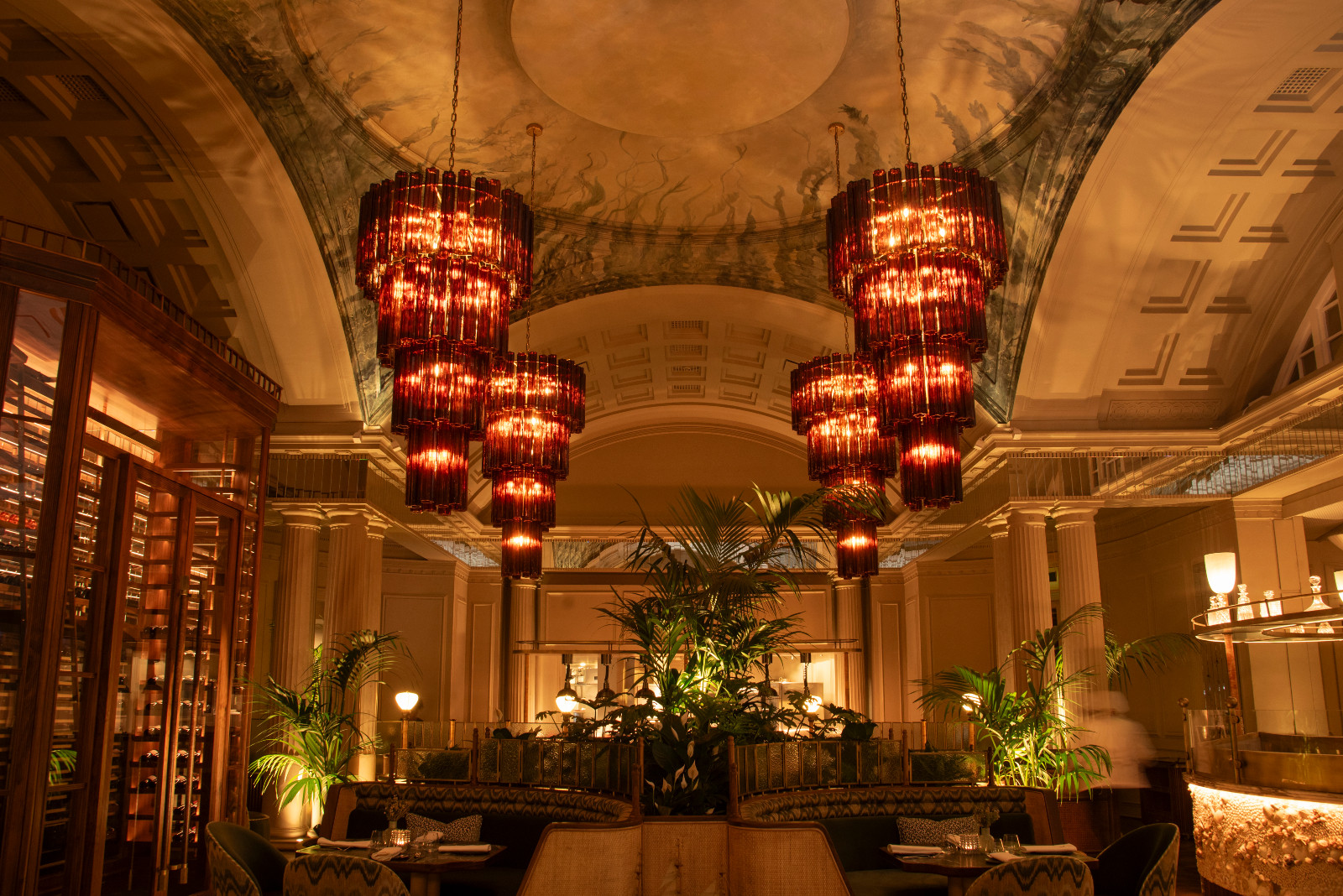 A wide view of Amura’s main dining room after dark, with four clusters of tiered amber glass chandeliers hanging from the painted vaulted ceiling, classical columns, a central planting arrangement and the wine cabinet to the left (Photo: Claire Gunn)