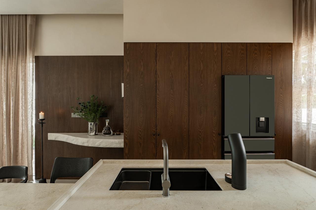 The kitchen island in limestone with a black undermount sink, looking towards the dark oak joinery wall and refrigerator integrated into the cabinetry run
