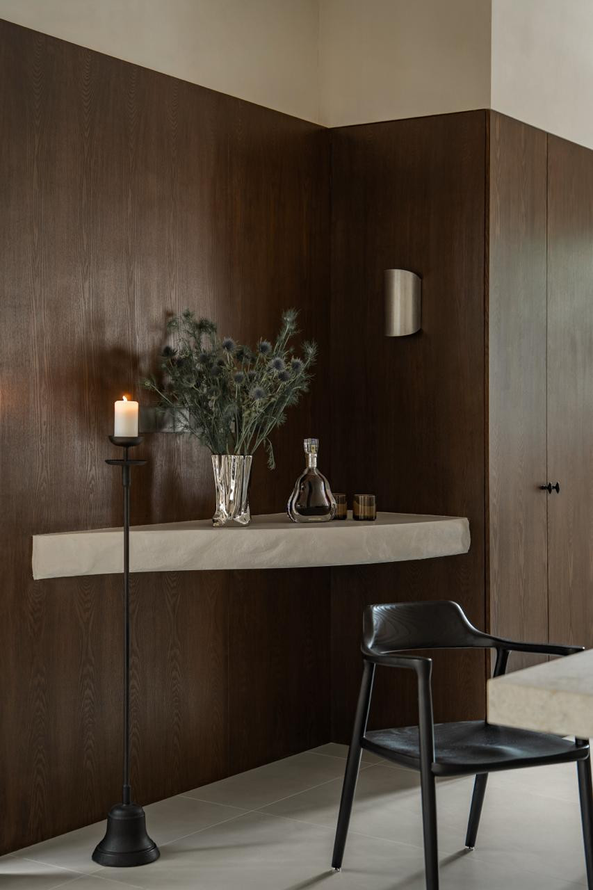 A floating stone console shelf against dark oak panelling, styled with dried thistles in a faceted silver vase, a glass decanter, and a black iron candle stand
