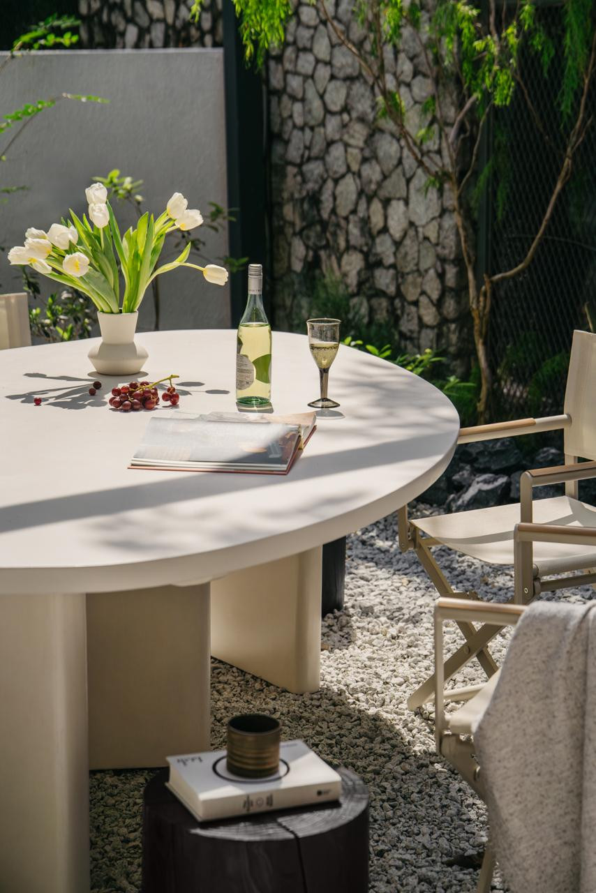 The oval dining table and canvas director’s chairs occupy the gravel courtyard, backed by a dry-stacked stone wall and dense tropical planting