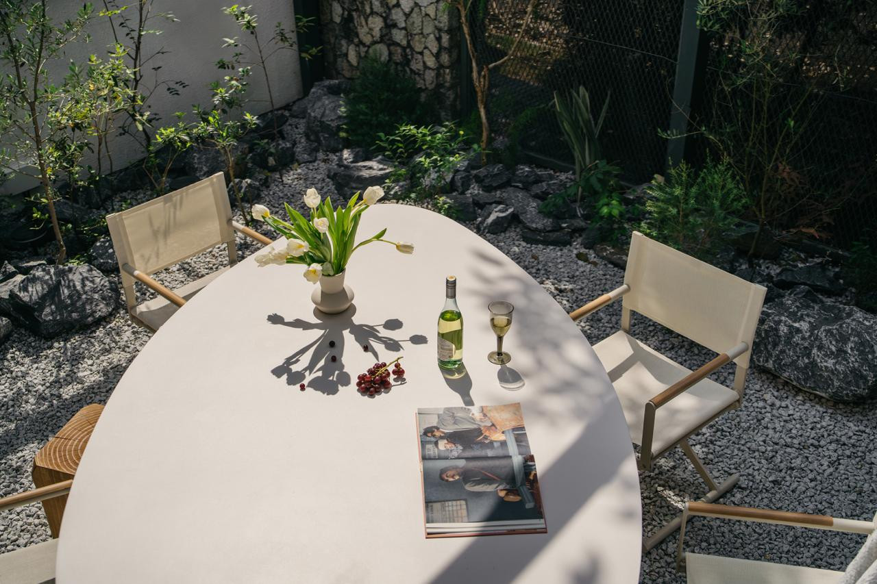 The oval outdoor dining table, set on white gravel among rocks and greenery, seen from above with tulips, a wine bottle, and an open magazine