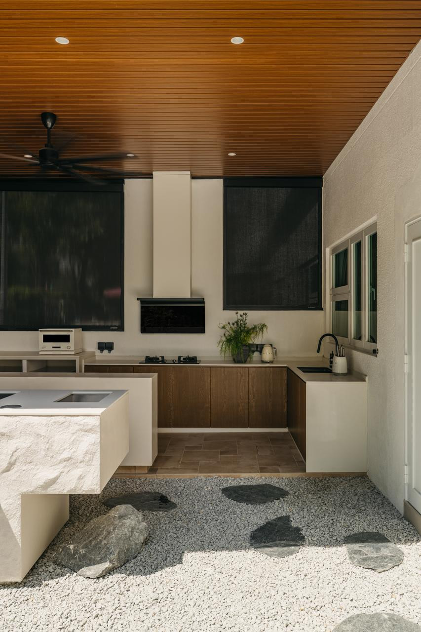 The covered outdoor kitchen features timber-lined ceilings, dark oak cabinetry, and stone countertops, opening directly onto the gravel courtyard below