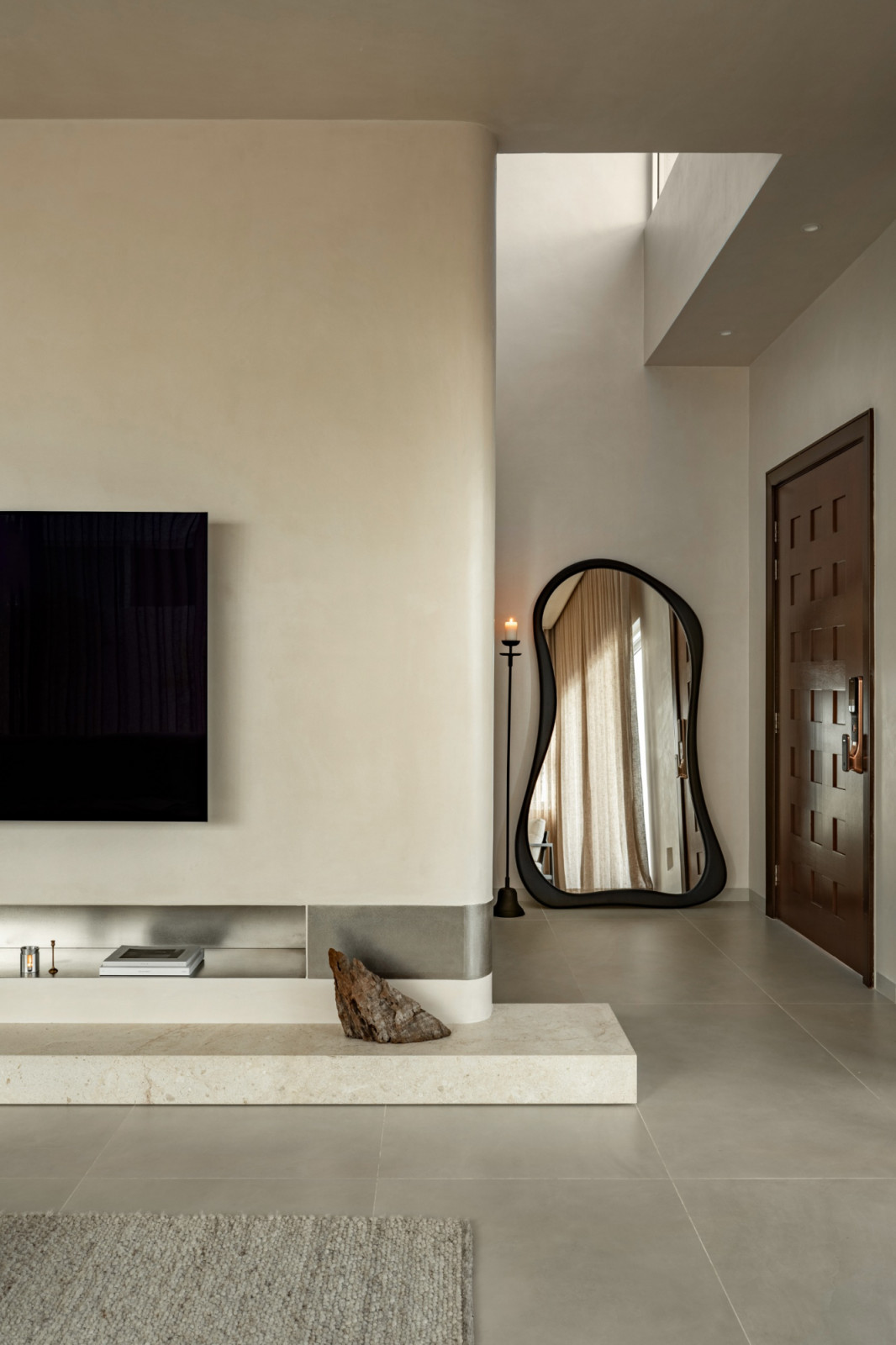 The living room TV wall in relation to the entry hall, where the organic-framed floor mirror and carved timber front door are visible; a skylight above the stairwell brings in natural light
