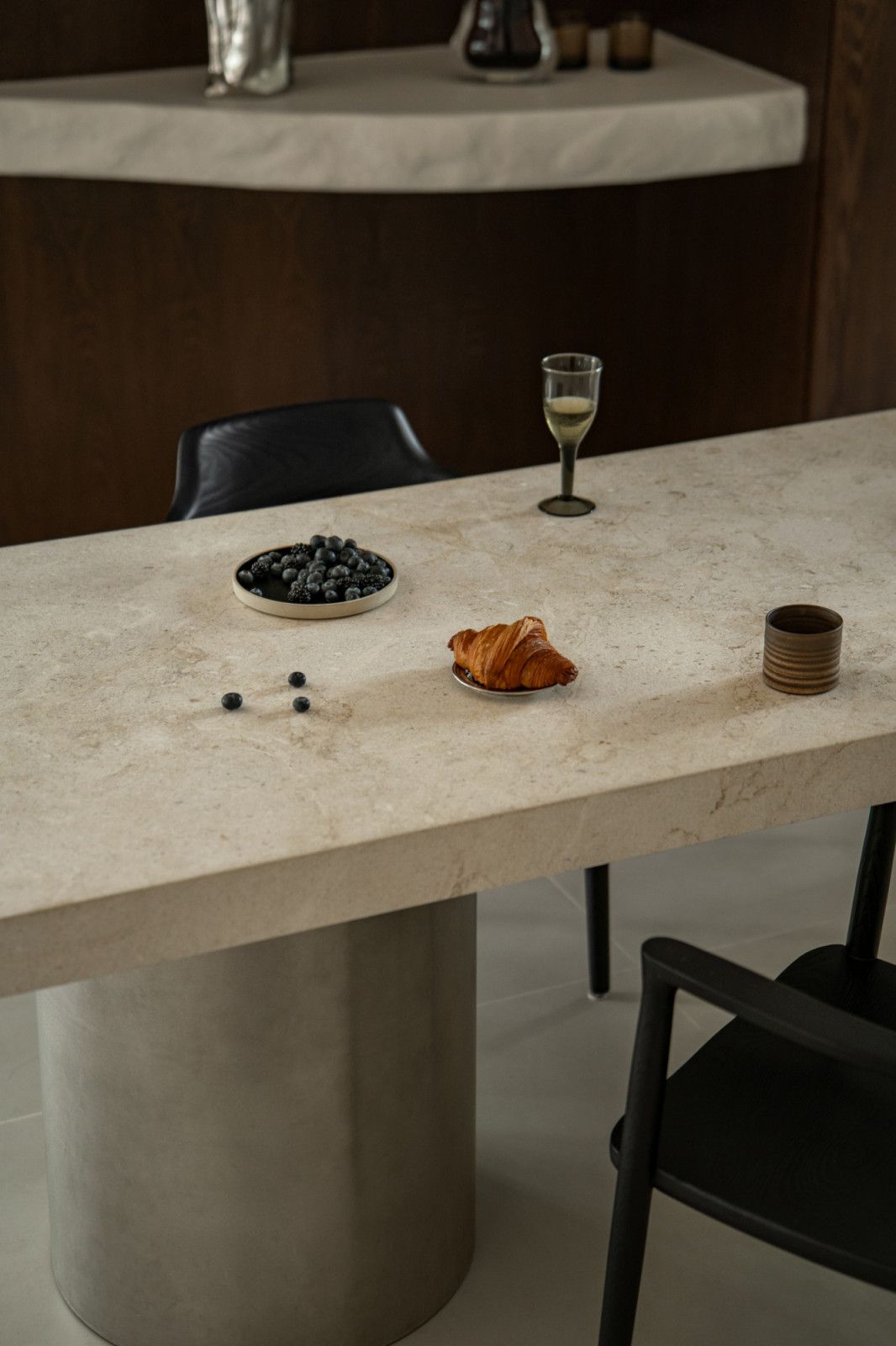 The limestone dining table surface, set with blueberries, a croissant, and a ceramic cup; the floating stone console and dark oak wall panels form the backdrop