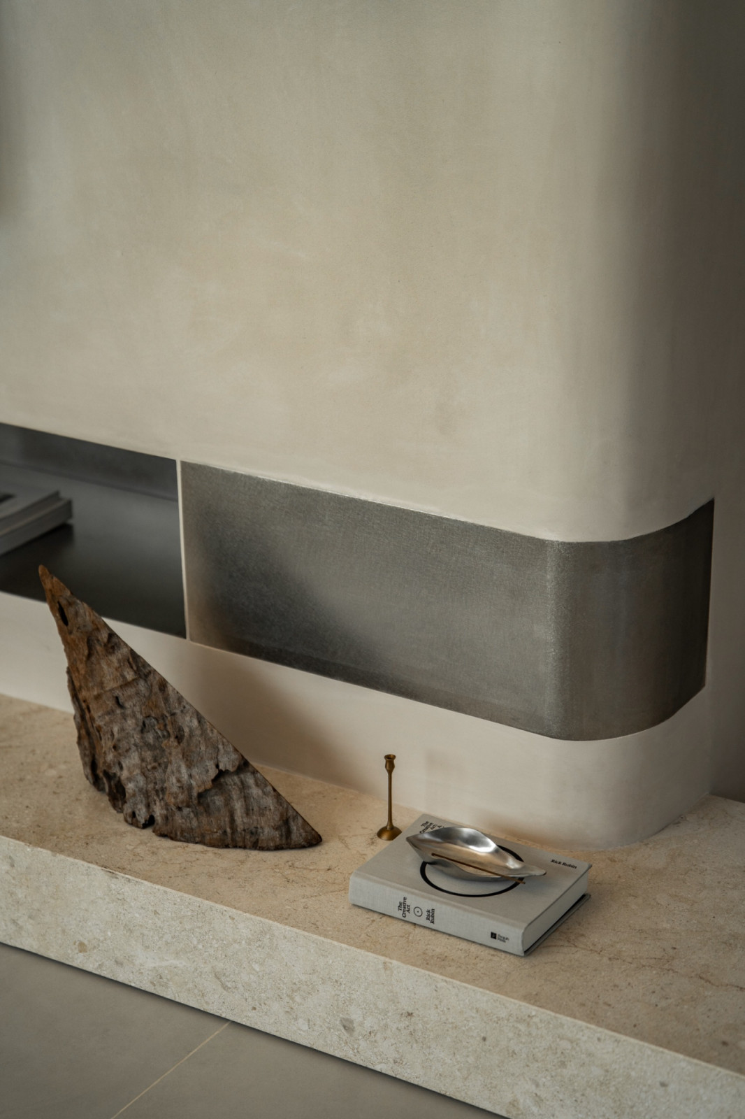 Close detail of the limestone TV plinth: a recessed brushed steel ledge, a piece of raw driftwood, a small brass candlestick, and a design book styled with a chrome object