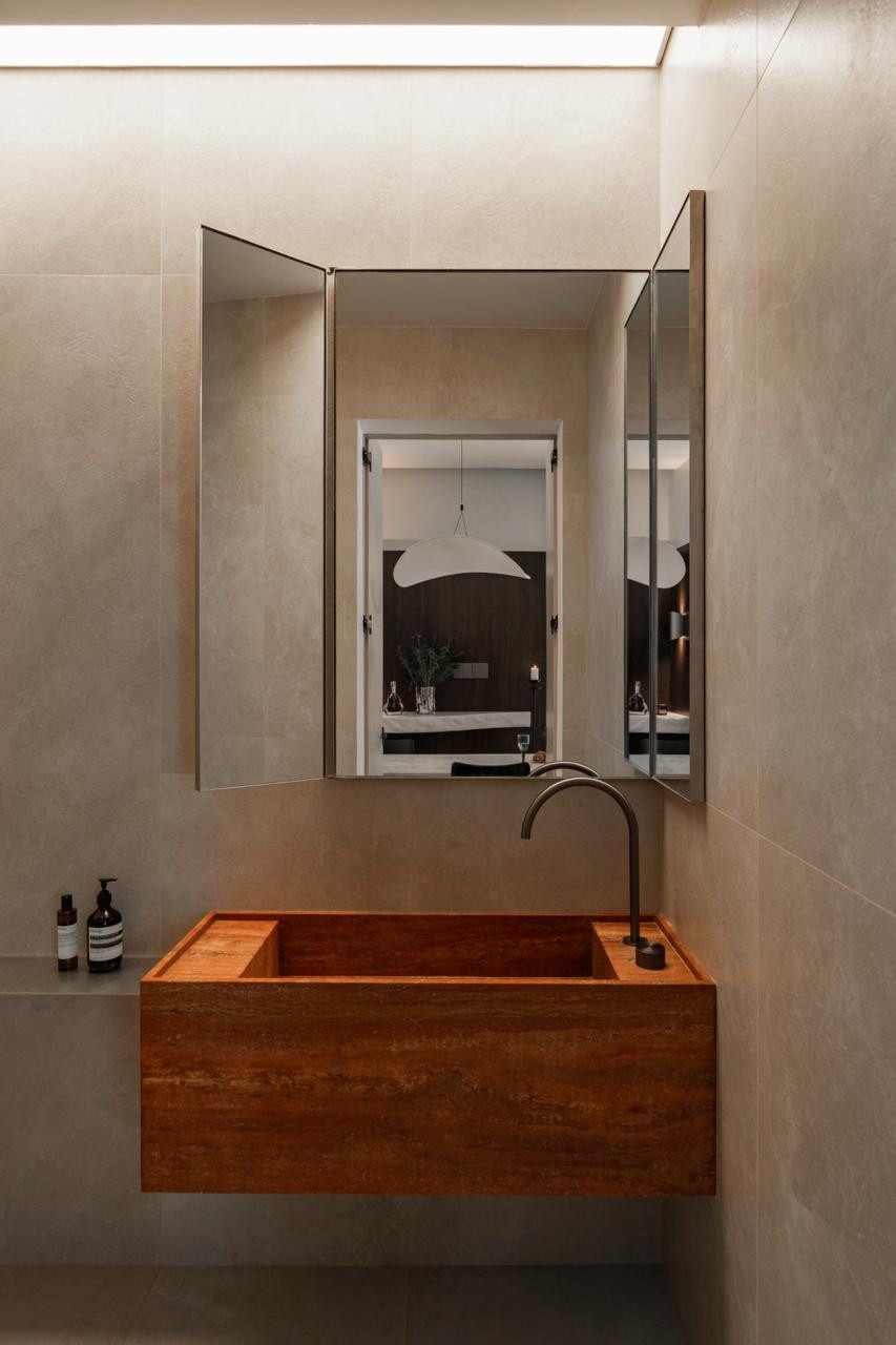 A box-form basin in warm terracotta-toned stone, paired with a curved gunmetal tap and a triptych mirror cabinet; concealed lighting runs along the ceiling edge